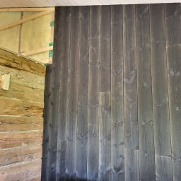 Usługi ogólnobudowlane Artur Klecha - Wnętrze domu w trakcie budowy: fragment ściany z bali drewnianych po lewej stronie i pionowo ułożone deski pomalowane na ciemnoszaro po prawej, widoczne ocieplenie z wełny mineralnej i elementy...