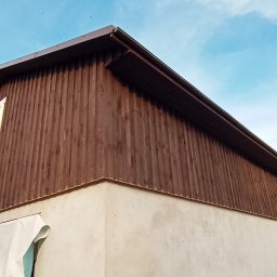 Usługi ogólnobudowlane Artur Klecha - Dwukondygnacyjny budynek z brązową, drewnianą elewacją na górnej części i otynkowaną, jasną dolną częścią, widoczne okno z szarą ramą i fragment zabezpieczającej folii budowlanej.