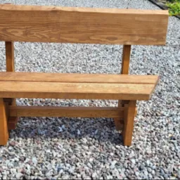 Drewniana ławka ogrodowa z oparciem, wykonana z surowego drewna, stojąca na żwirowym podłożu. W tle widoczne ułożone deski i zieleń.