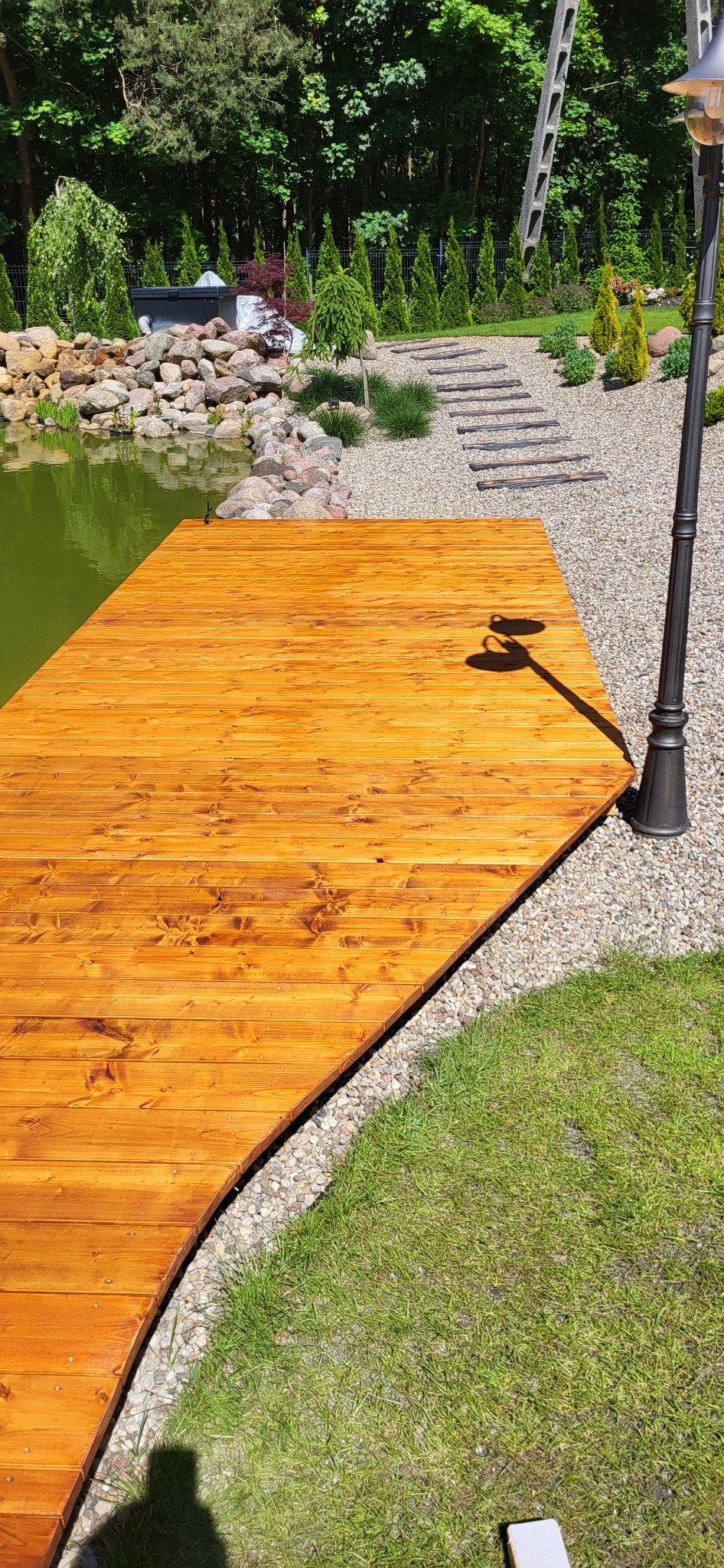 Nowoczesny pomost z desek o ciepłym, miodowym odcieniu, przylegający do zielonego stawu, z widocznym oświetleniem ogrodowym i ścieżką z kamieni prowadzącą do ogrodu.