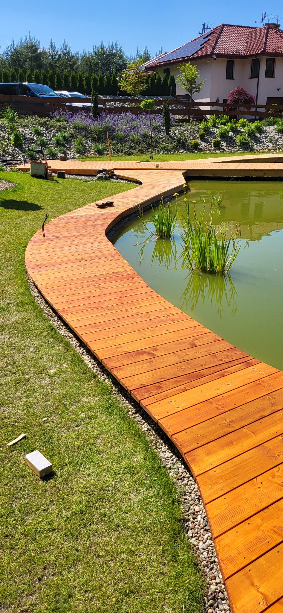 Zakrzywiona drewniana ścieżka nad stawem, wykonana z desek w ciepłym odcieniu, otoczona zielonym trawnikiem i kamieniami, prowadząca do dalszej części ogrodu z roślinnością i budynkiem w tle.