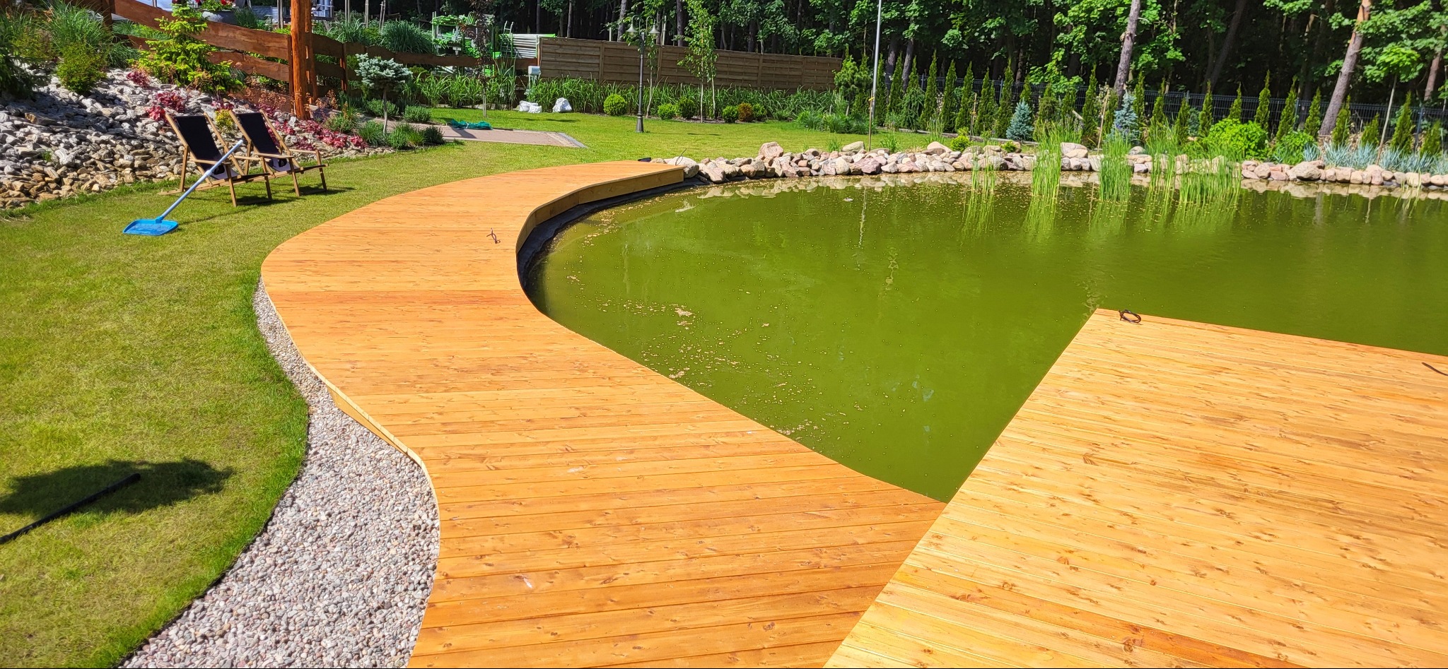 Zakrzywiony pomost z jasnego drewna nad zielonkawym stawem, otoczony kamieniami i trawnikiem, z dwoma krzesłami plażowymi i niebieską szufelką w tle.