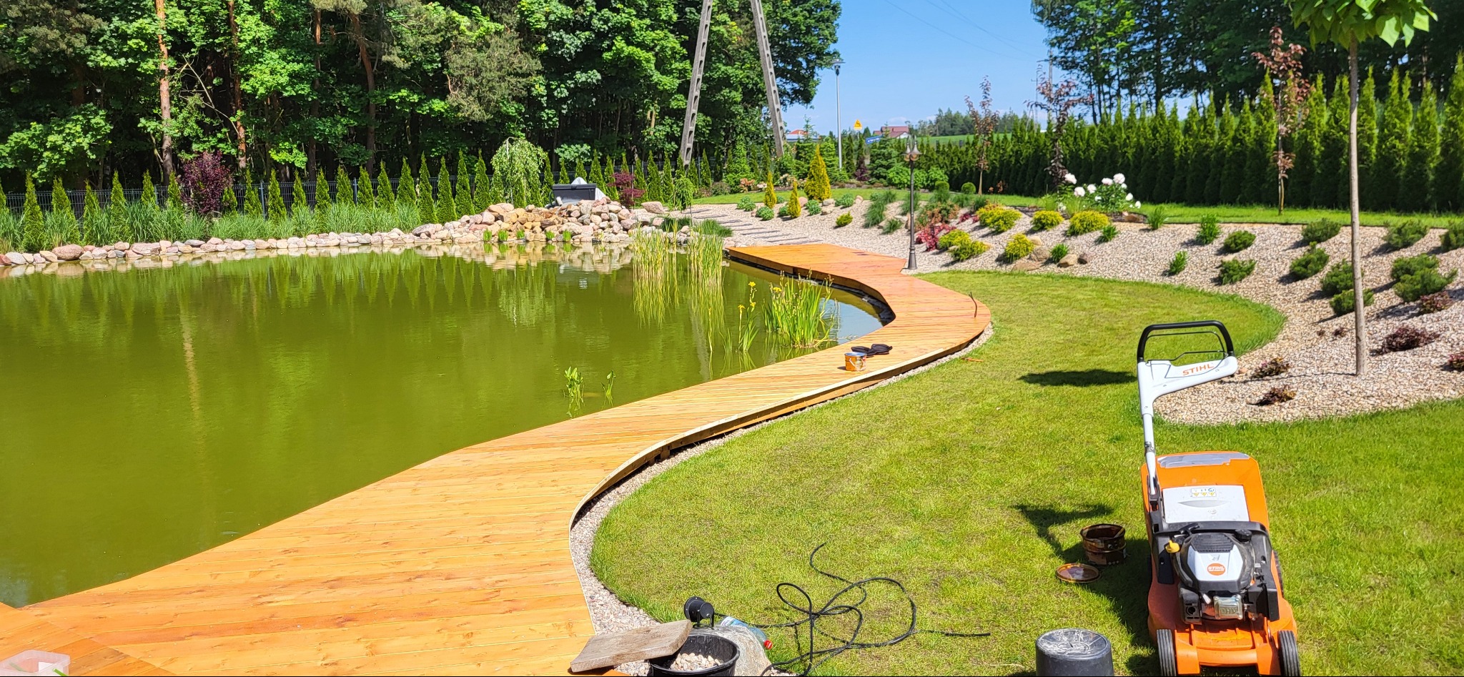Widok na zadbany ogród z oczkiem wodnym i wijącym się drewnianym pomostem, kosiarka do trawy stoi na trawniku, w tle liczne rośliny ozdobne i drzewa.