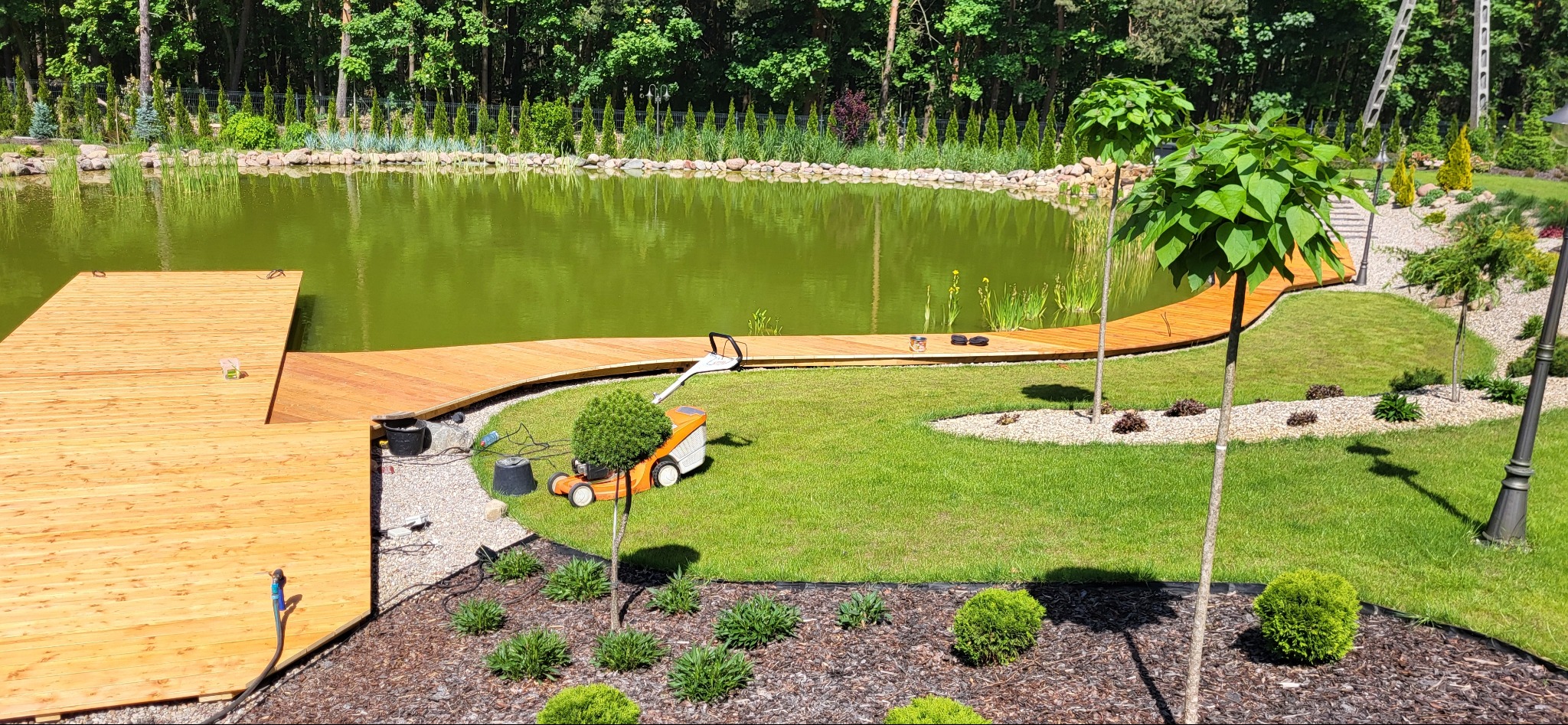 Widok z góry na drewniany pomost nad stawem, z zakrzywioną ścieżką, kosiarką do trawy, młodymi drzewkami i ozdobnymi krzewami w zadbanym ogrodzie.