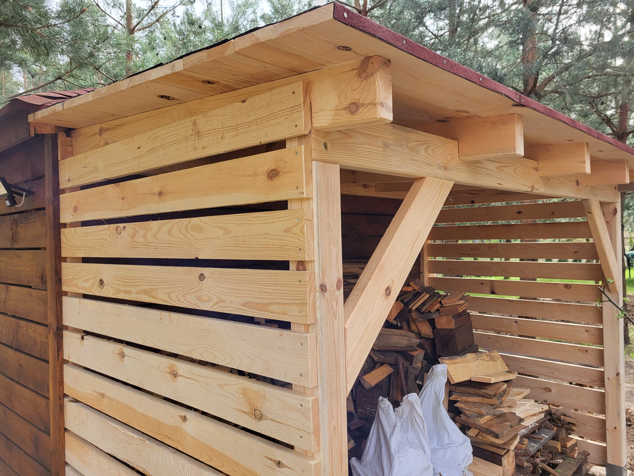 Drewniana konstrukcja z dachem krytym papą, prawdopodobnie wiata na drewno, wypełniona pociętymi kawałkami drewna i zabezpieczona białym workiem, widoczna na tle drzew.