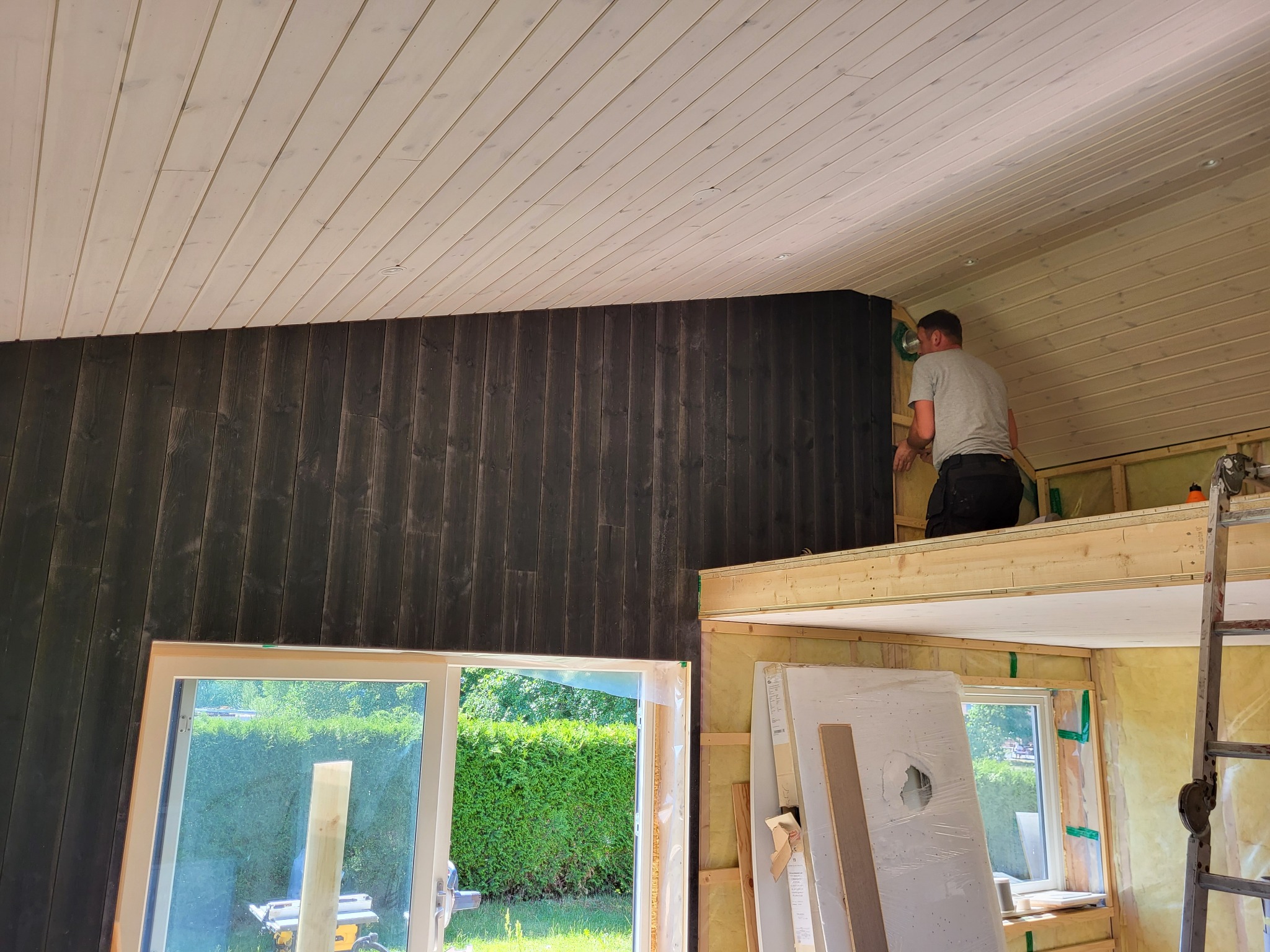 Wnętrze domu w trakcie budowy, widoczna antresola z drewnianą konstrukcją, ściany wykończone ciemnym drewnem, biały sufit z drewnianych paneli, okno z widokiem na zieleń, drabina i materiały...