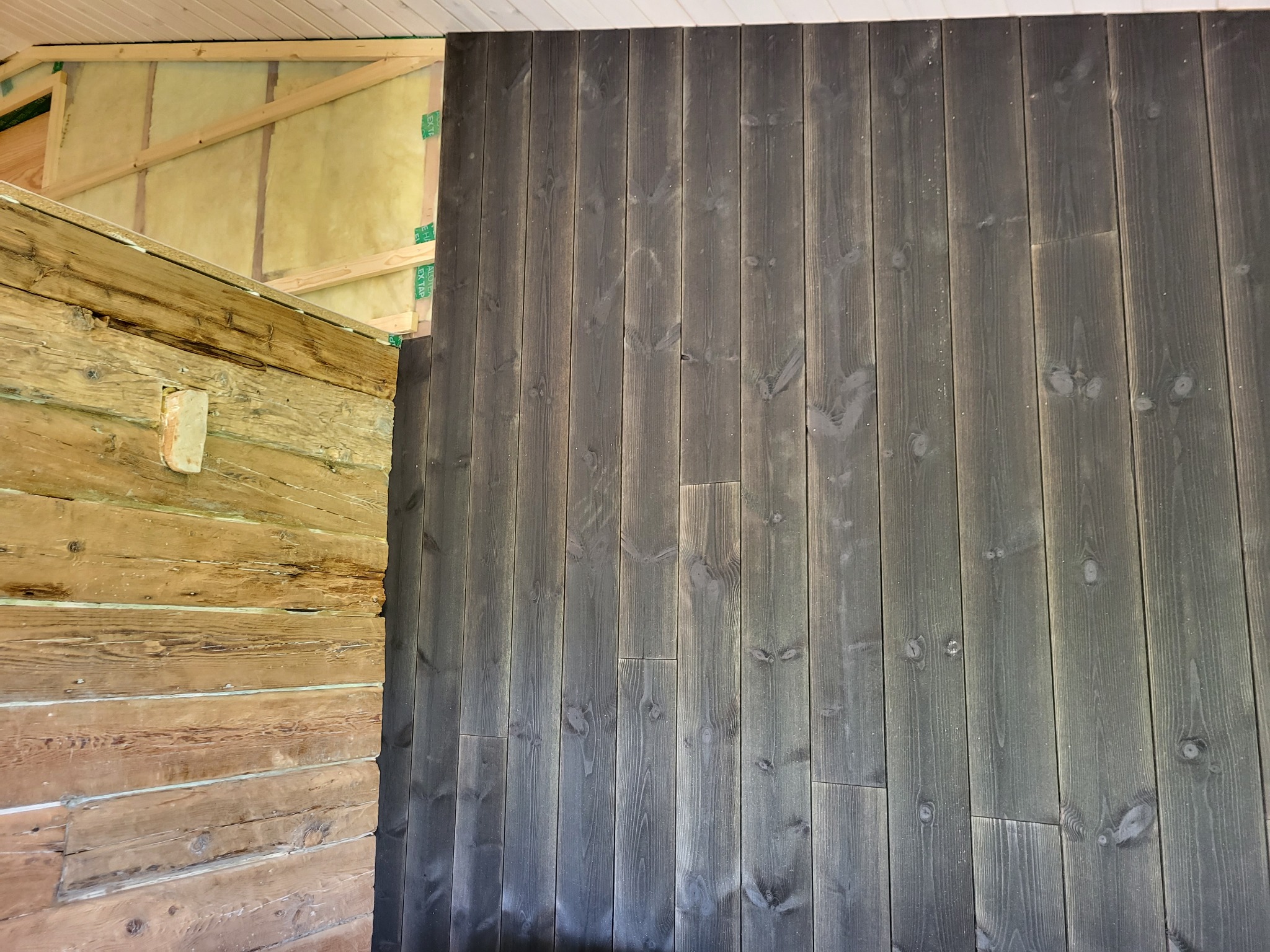 Wnętrze domu w trakcie budowy: fragment ściany z bali drewnianych po lewej stronie i pionowo ułożone deski pomalowane na ciemnoszaro po prawej, widoczne ocieplenie z wełny mineralnej i elementy...