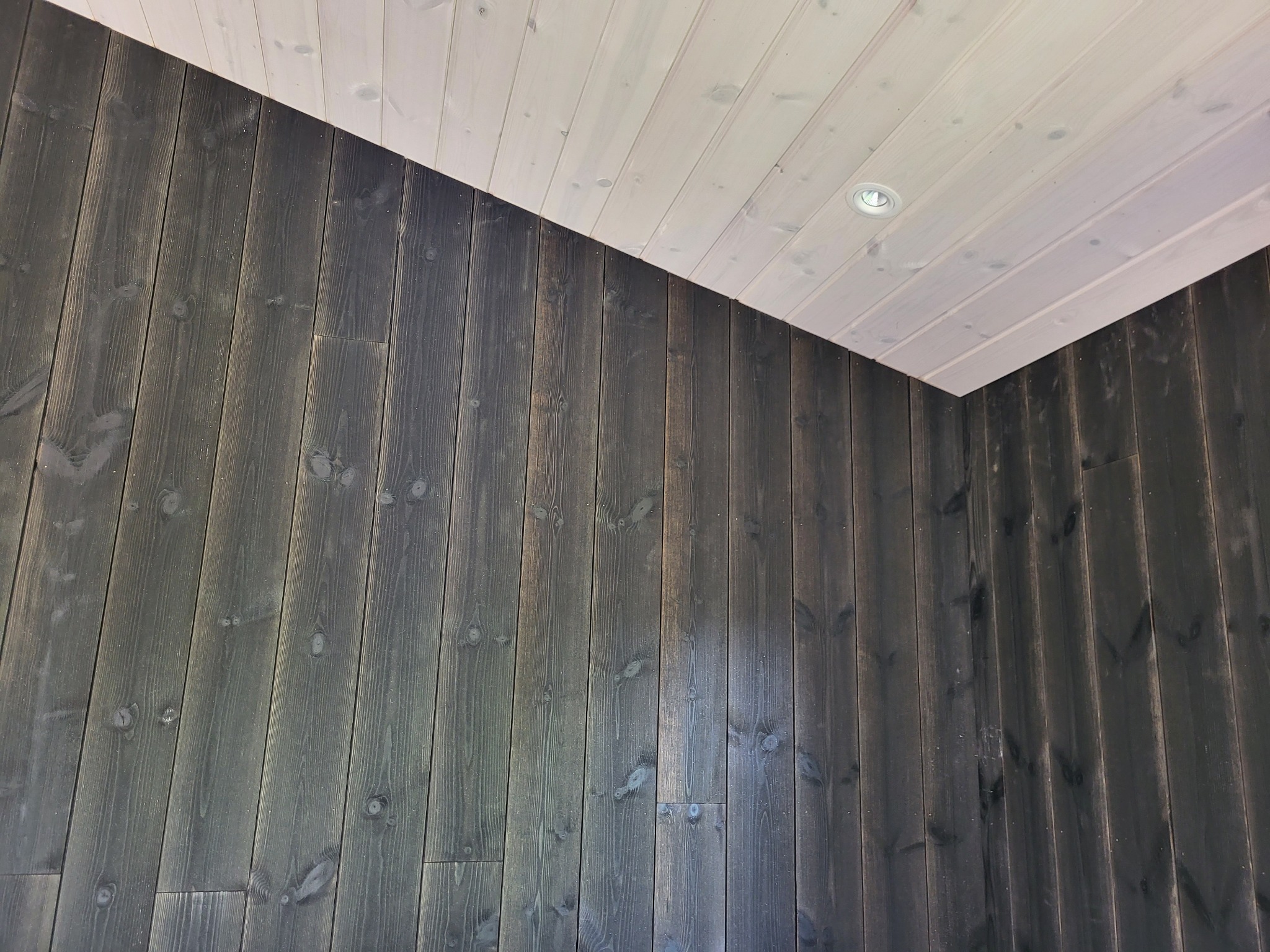 Wnętrze z ciemną, pionowo ułożoną boazerią i jasnym, drewnianym sufitem z wbudowanym oświetleniem punktowym.