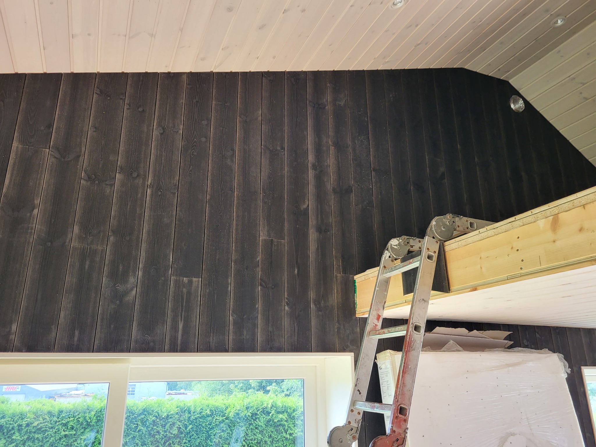 Wnętrze w trakcie wykańczania: ściana obita ciemnym drewnem, biały sufit z desek, okno z widokiem na zieleń, drabina oparta o konstrukcję antresoli z materiałami budowlanymi w folii.