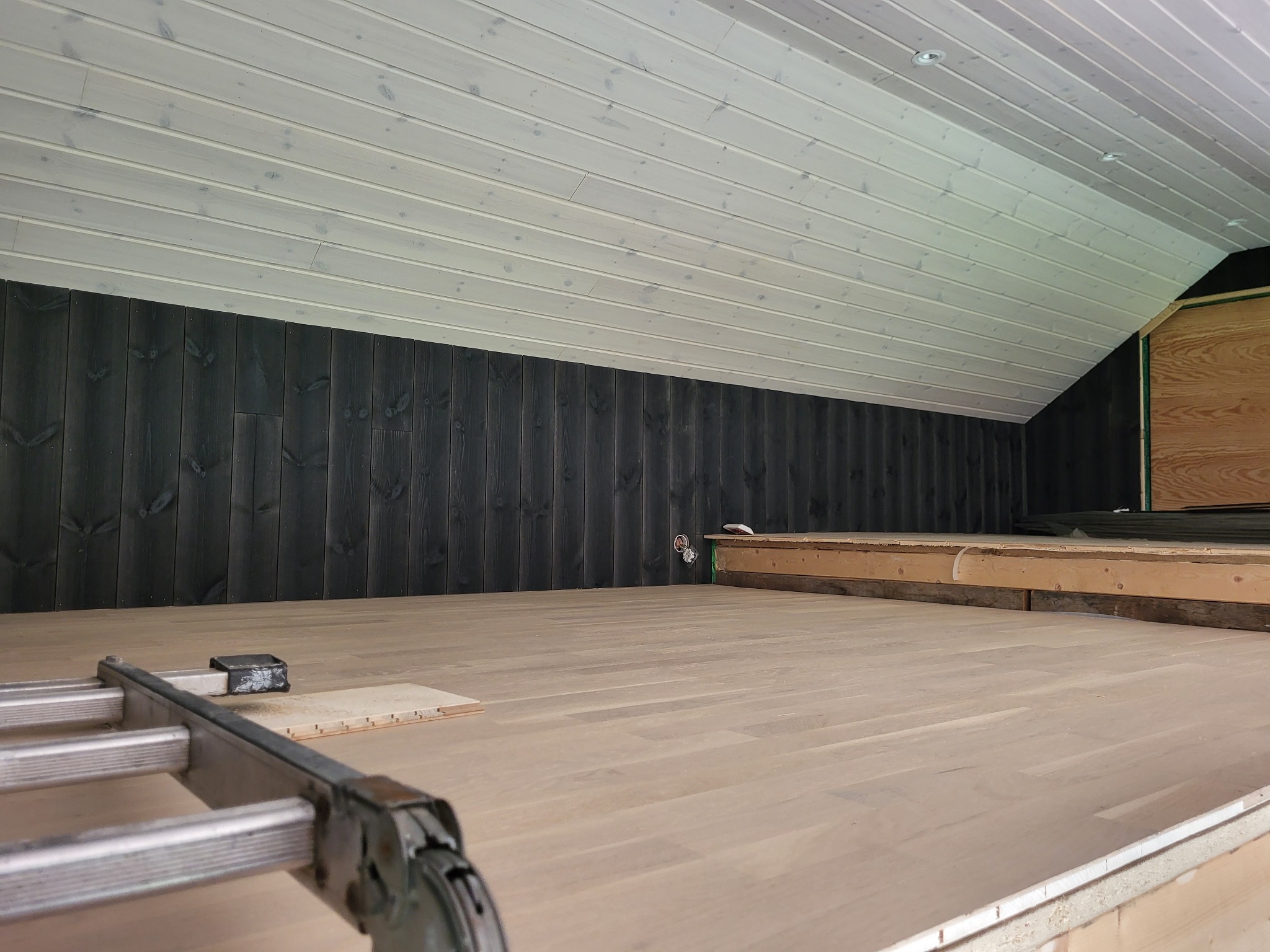 Wnętrze poddasza w trakcie adaptacji: widoczne drewniane panele podłogowe, czarne deski na ścianie i białe deski na skośnym suficie, drabina w rogu pomieszczenia.