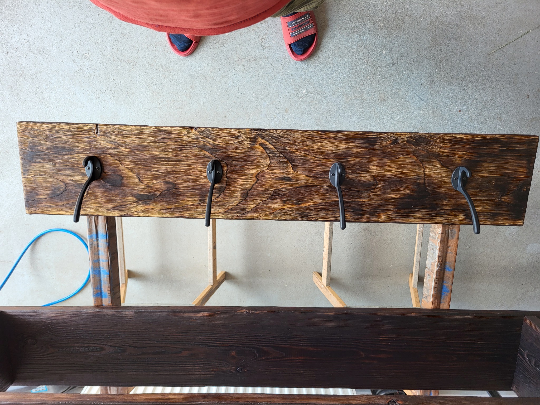 Wieszak ścienny z ciemnego drewna z czterema czarnymi haczykami, przymocowany do prowizorycznego stojaka podczas prac wykończeniowych, widok z góry.