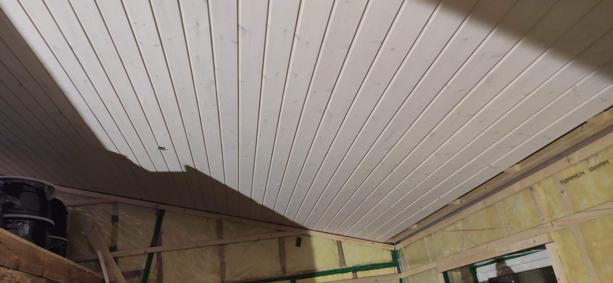 Wnętrze domu w trakcie budowy, widoczny sufit z jasnych desek boazeryjnych, ocieplenie ścian wełną mineralną i drewniana konstrukcja szkieletowa.