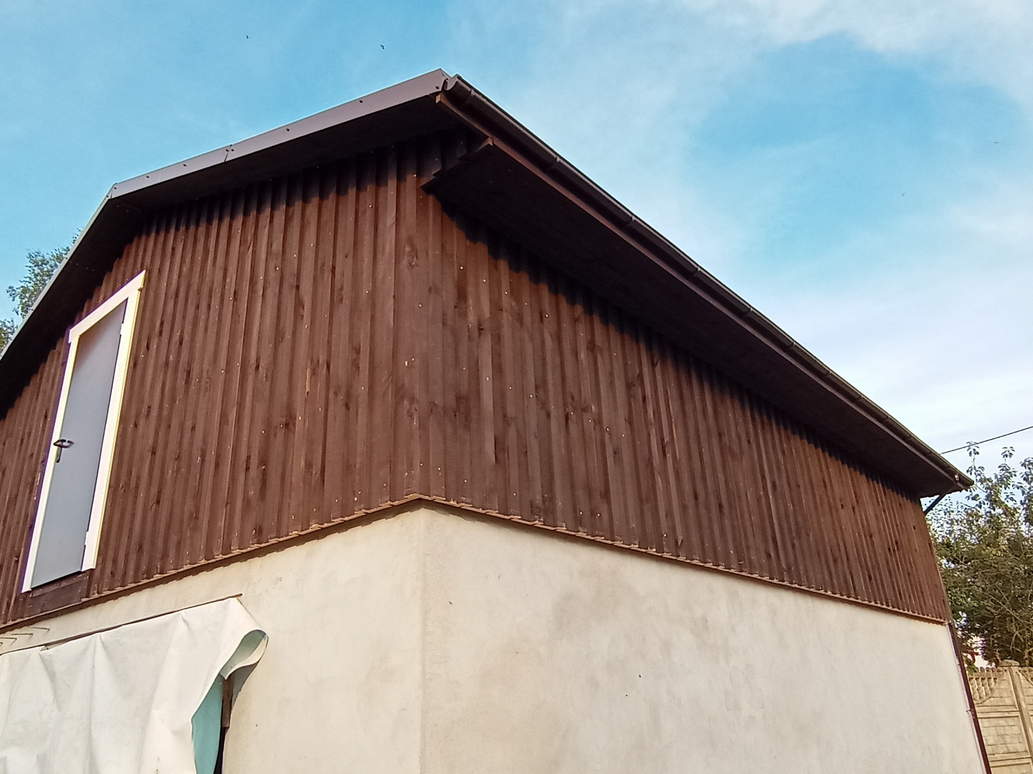 Dwukondygnacyjny budynek z brązową, drewnianą elewacją na górnej części i otynkowaną, jasną dolną częścią, widoczne okno z szarą ramą i fragment zabezpieczającej folii budowlanej.