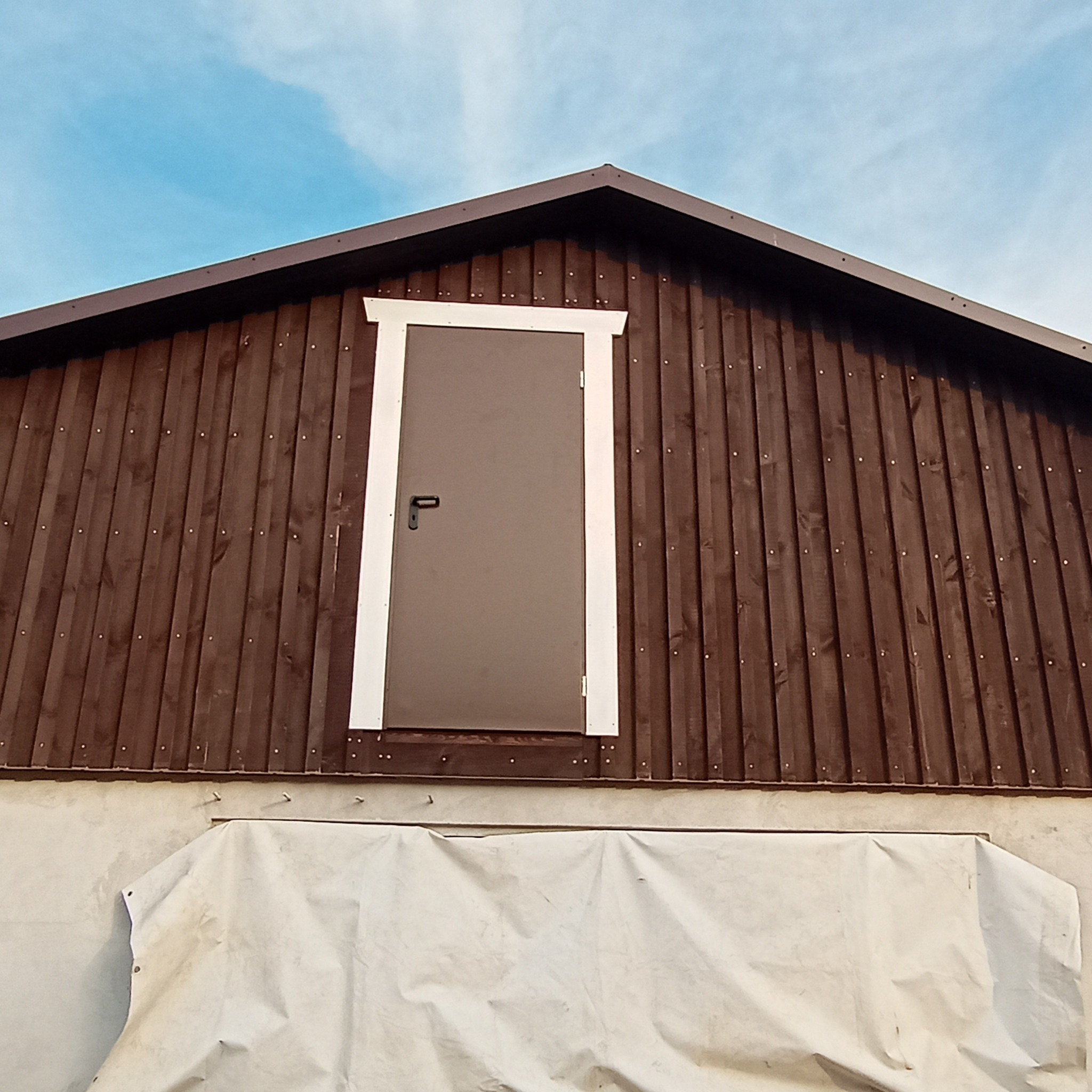 Brązowa brama w szczycie budynku z drewnianą fasadą, obramowana białą listwą, zabezpieczona plandeką na dole, widok z dołu.