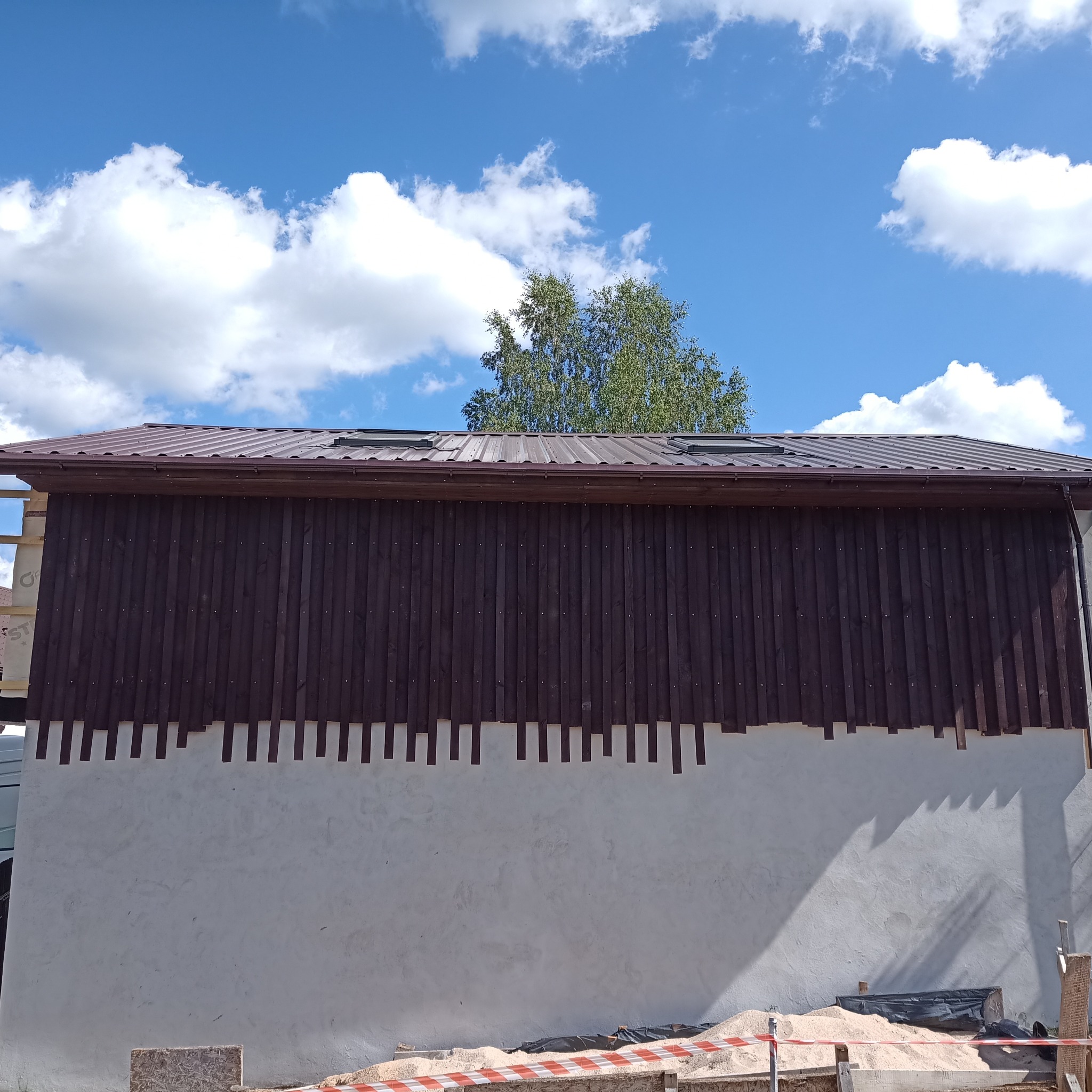 Ściana budynku w trakcie montażu brązowej okładziny pionowej, widoczne fragmenty rusztowania, dach z brązowej blachy i błękitne niebo z chmurami.