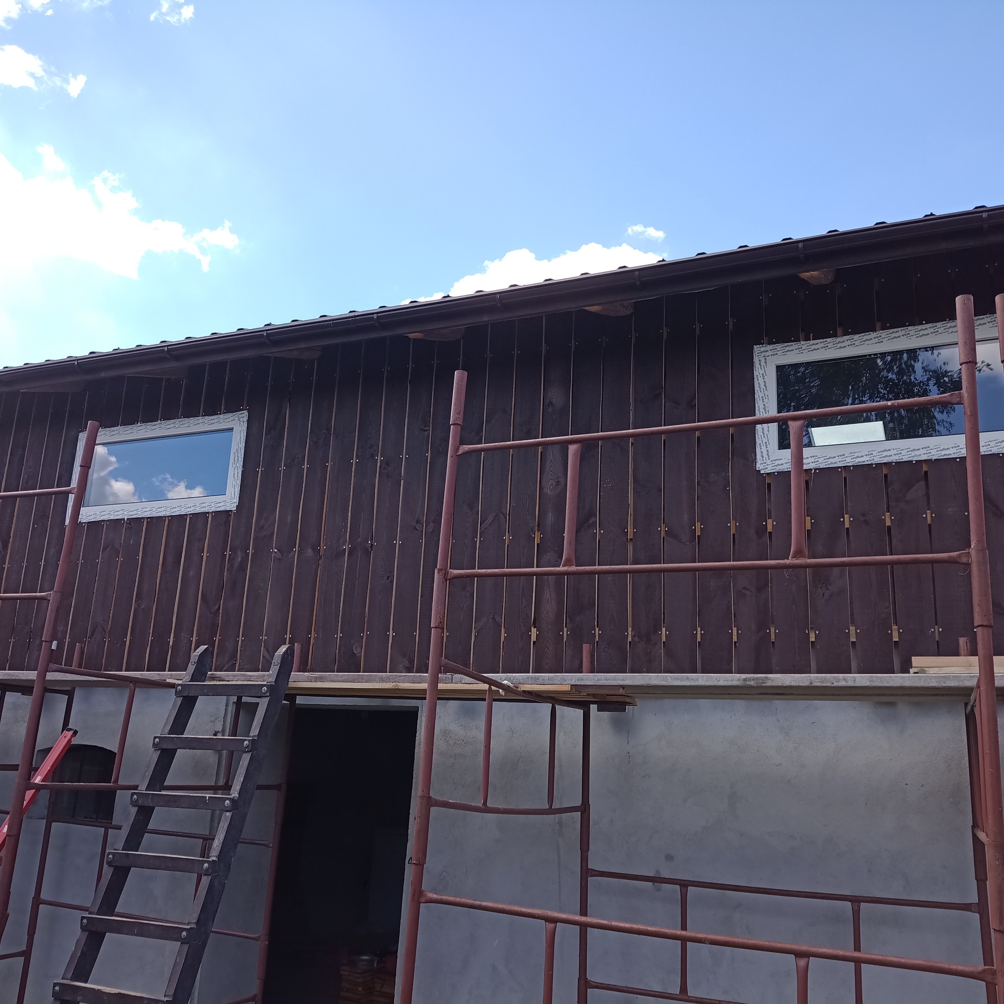 Budynek w trakcie montażu drewnianej elewacji, widoczne rusztowanie i drabina, okna z białymi ramami, pionowe deski w ciemnobrązowym kolorze.