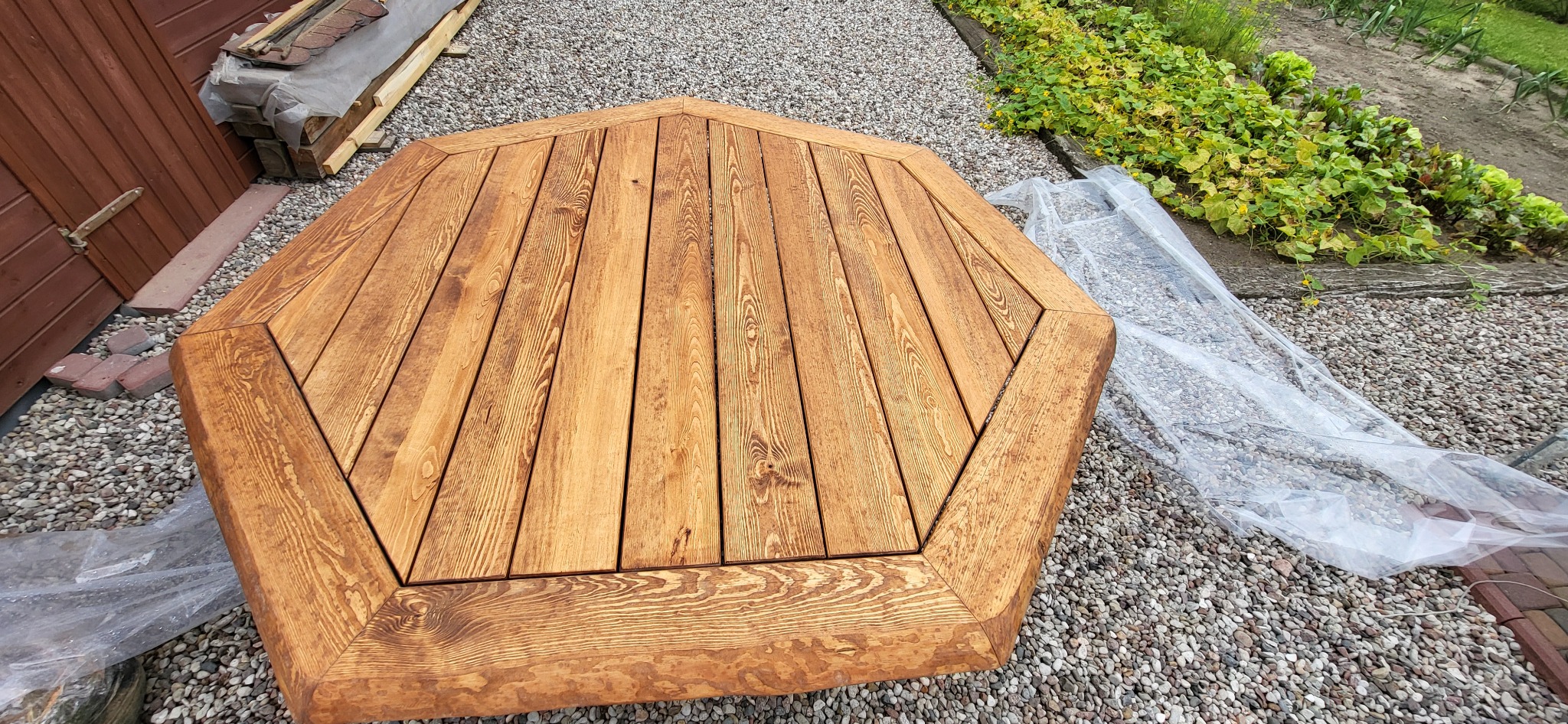 Ośmiokątny, impregnowany stół z drewna na zewnątrz, na żwirowym podłożu, częściowo przykryty folią ochronną, widoczne elementy konstrukcyjne budynku w tle.