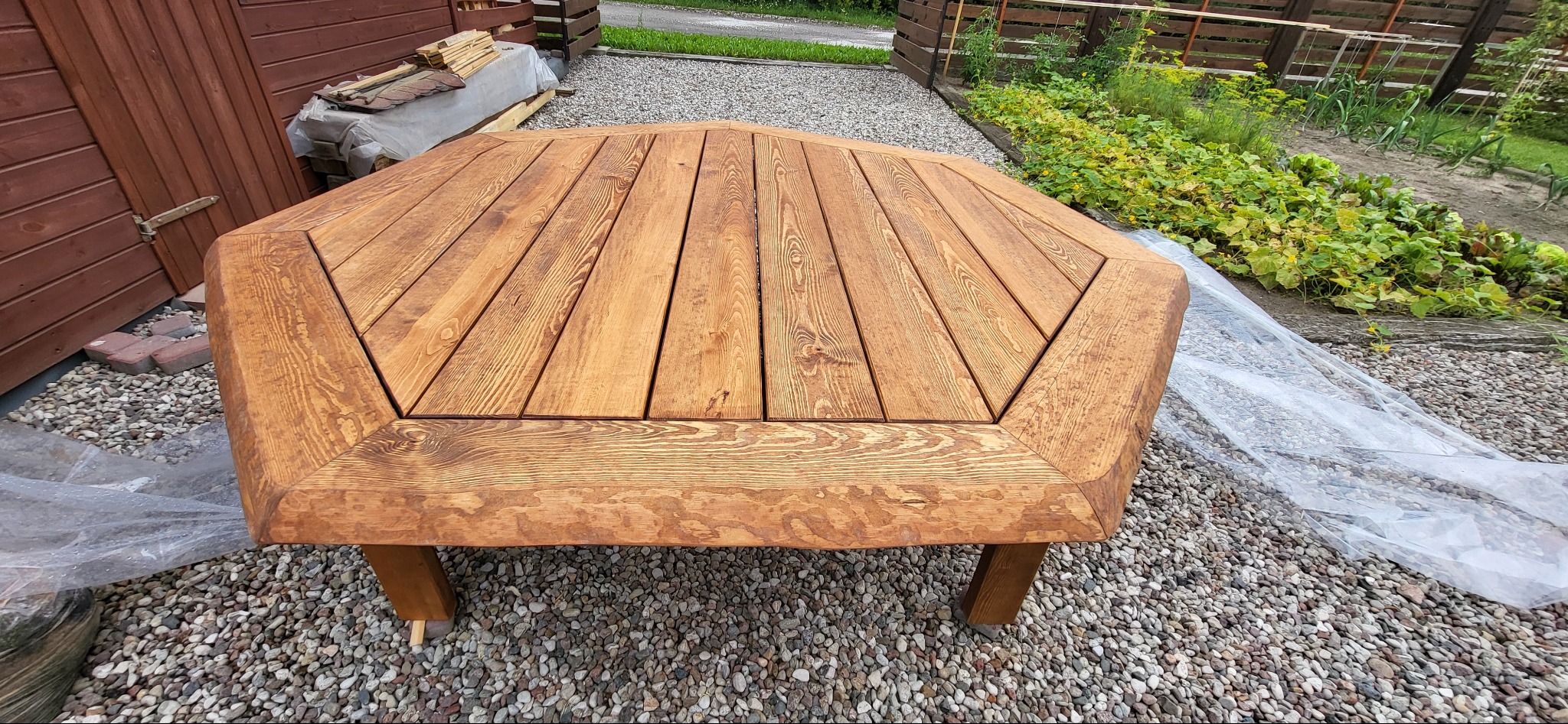 Stół ogrodowy z drewna o nietypowym, wielokątnym kształcie, wykonany przez stolarza z Dąbrówki, ustawiony na żwirowym podłożu, z widocznym ogrodem w tle.