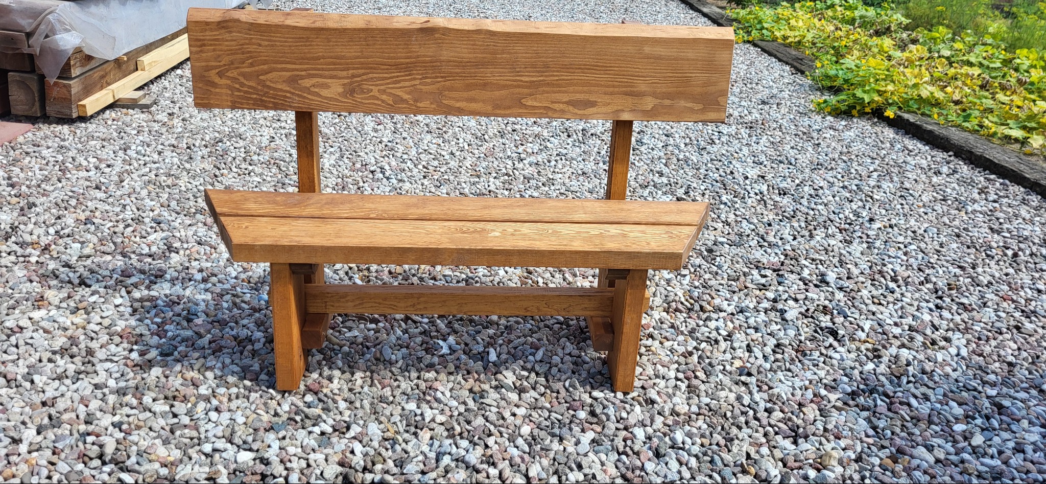 Drewniana ławka ogrodowa z oparciem, wykonana z surowego drewna, stojąca na żwirowym podłożu. W tle widoczne ułożone deski i zieleń.