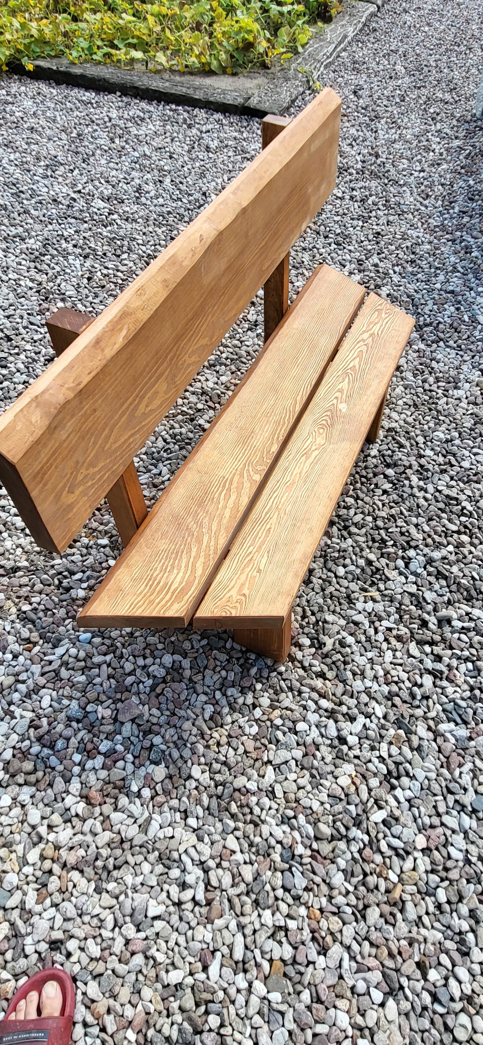 Drewniana ławka ogrodowa z oparciem, wykonana z jasnego drewna, ustawiona na żwirowym podłożu. Widoczny fragment stopy w klapku w lewym dolnym rogu.