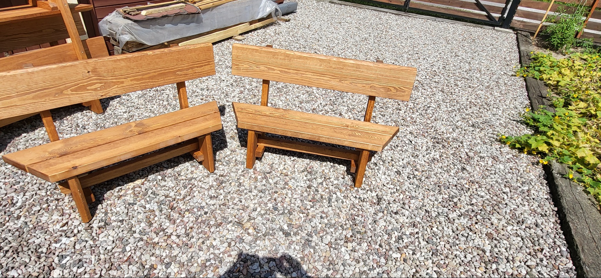Dwie nowe, drewniane ławki z oparciami stoją na żwirowym podłożu w ogrodzie, widoczne słoje drewna i naturalny kolor.