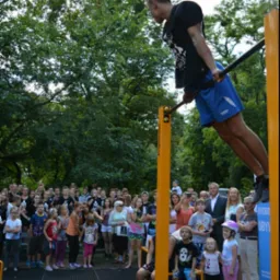 Mężczyzna wykonuje ćwiczenie siłowe na drążku, trzymając się go rękami i unosząc ciało, w tle obserwujący tłum ludzi i drzewa w parku.