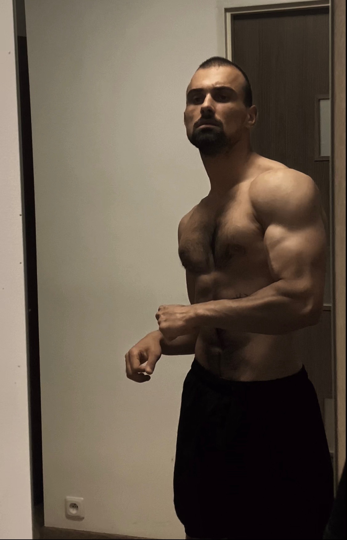 Mężczyzna z wyraźnie zarysowanymi mięśniami, pozujący w czarnych spodenkach na tle białej ściany i częściowo widocznych drzwi