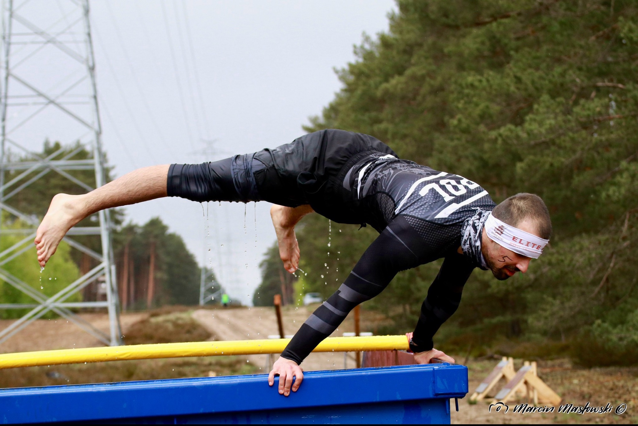 Mężczyzna w stroju sportowym, z opaską na głowie, pokonuje przeszkodę podczas biegu OCR, w tle las i słupy wysokiego napięcia.