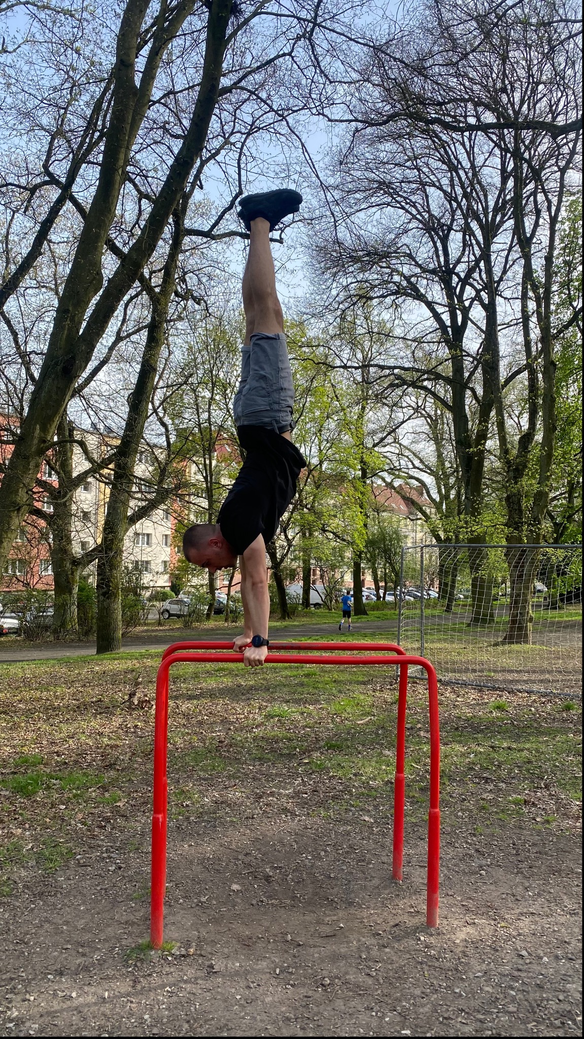Mężczyzna w czarnej koszulce i szarych spodenkach wykonuje stójkę na rękach na czerwonych poręczach na zewnątrz, w tle drzewa i budynki.