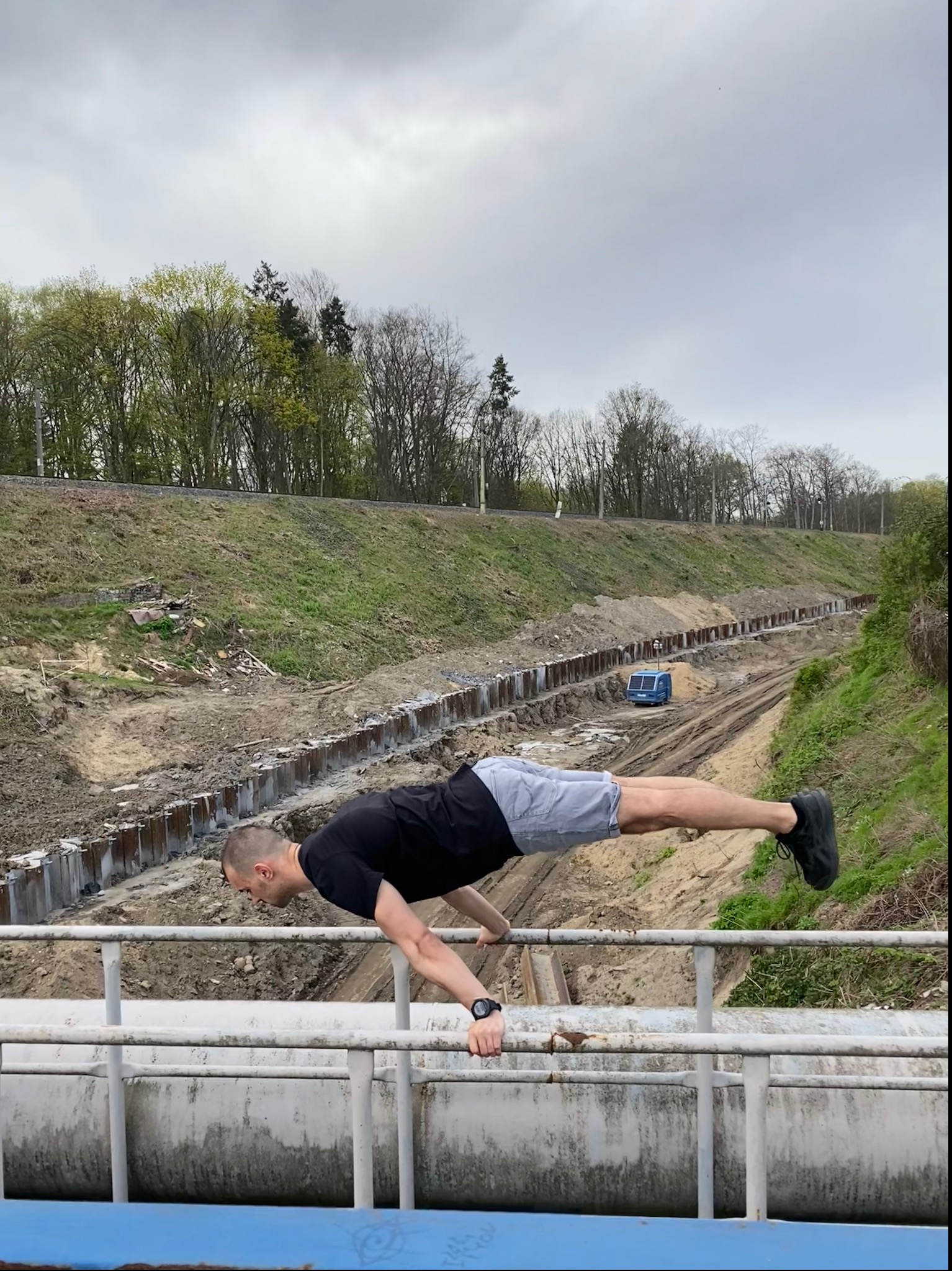 Mężczyzna w czarnej koszulce i szarych spodenkach wykonuje zaawansowaną pozycję plank na metalowej barierce, z widokiem na wykop i zarośnięte zbocze w tle.