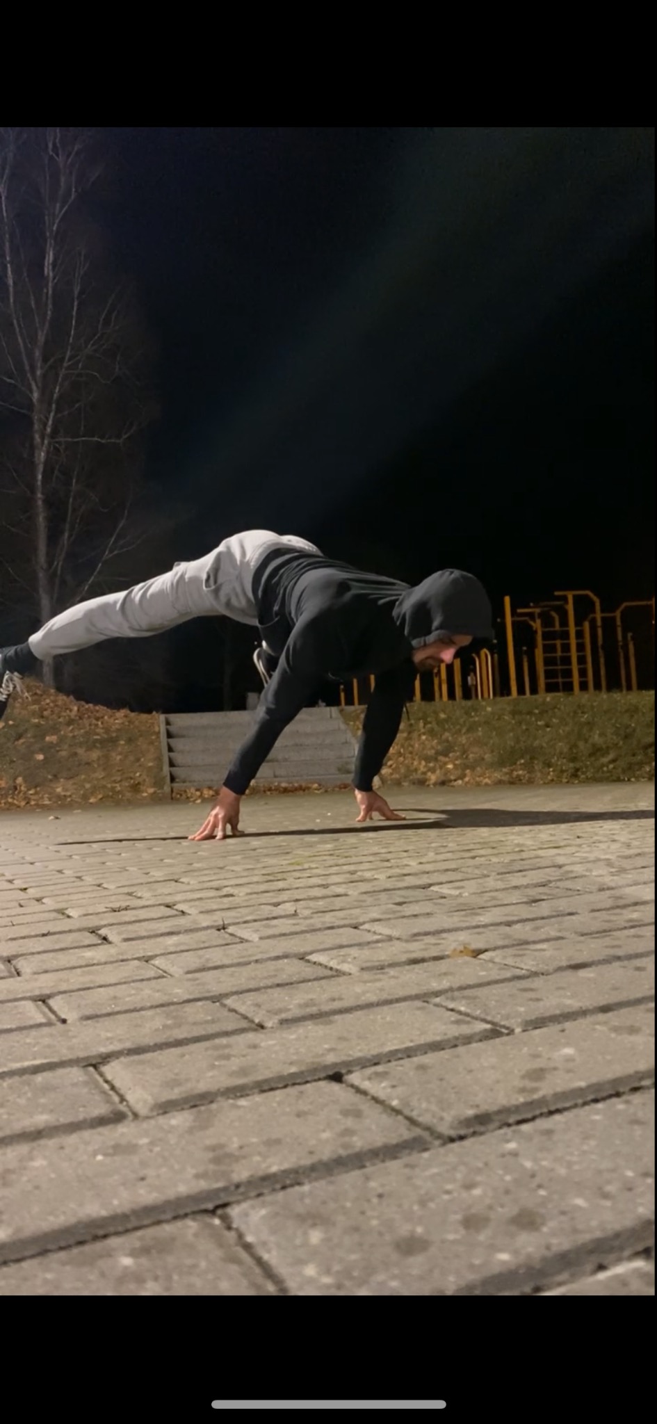 Mężczyzna w czarnej bluzie z kapturem wykonuje zaawansowaną pozycję kalisteniki na chodniku z kostki brukowej, w tle drzewo i elementy placu do ćwiczeń na świeżym powietrzu, nocne oświetlenie.