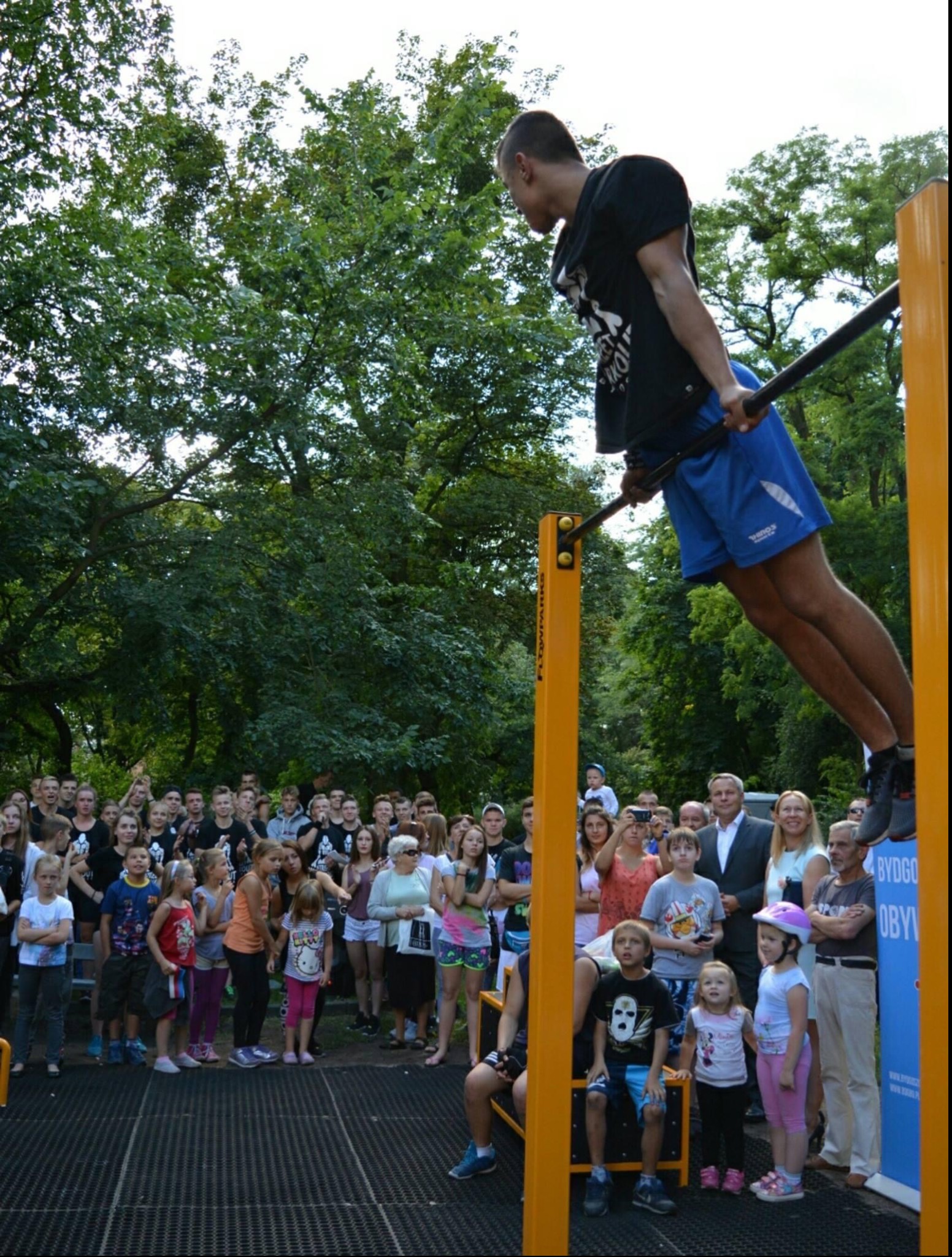 Mężczyzna wykonuje ćwiczenie siłowe na drążku, trzymając się go rękami i unosząc ciało, w tle obserwujący tłum ludzi i drzewa w parku.