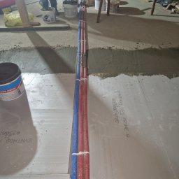 Jakub Markowski - Instalacja podłogowa: rury grzewcze, niebieskie i czerwone, ułożone w wykopie na izolacji podłogowej, widoczne narzędzia i wiaderka z zaprawą w tle.