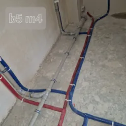 Nowe rury wodne i grzewcze, czerwone i niebieskie, prowadzone wzdłuż ściany i podłogi, zabezpieczone białymi opaskami zaciskowymi, w surowym wnętrzu.