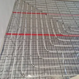 Białe rurki ogrzewania podłogowego, mocowane czarnymi klipsami do folii z nadrukowaną siatką, ułożone na izolacji ze srebrną folią aluminiową, z widocznymi czerwonymi pasami taśmy mocującej, przed...