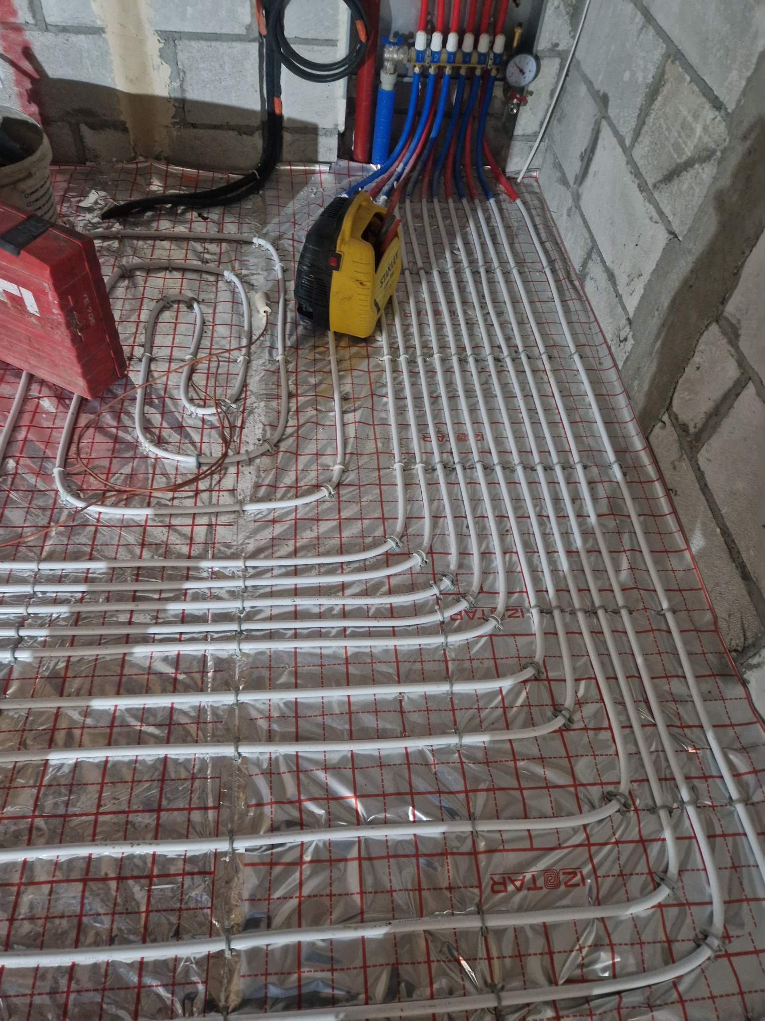 Białe rurki ogrzewania podłogowego zamocowane do siatki z folią aluminiową, widoczne narzędzia i elementy instalacji w surowym wnętrzu.