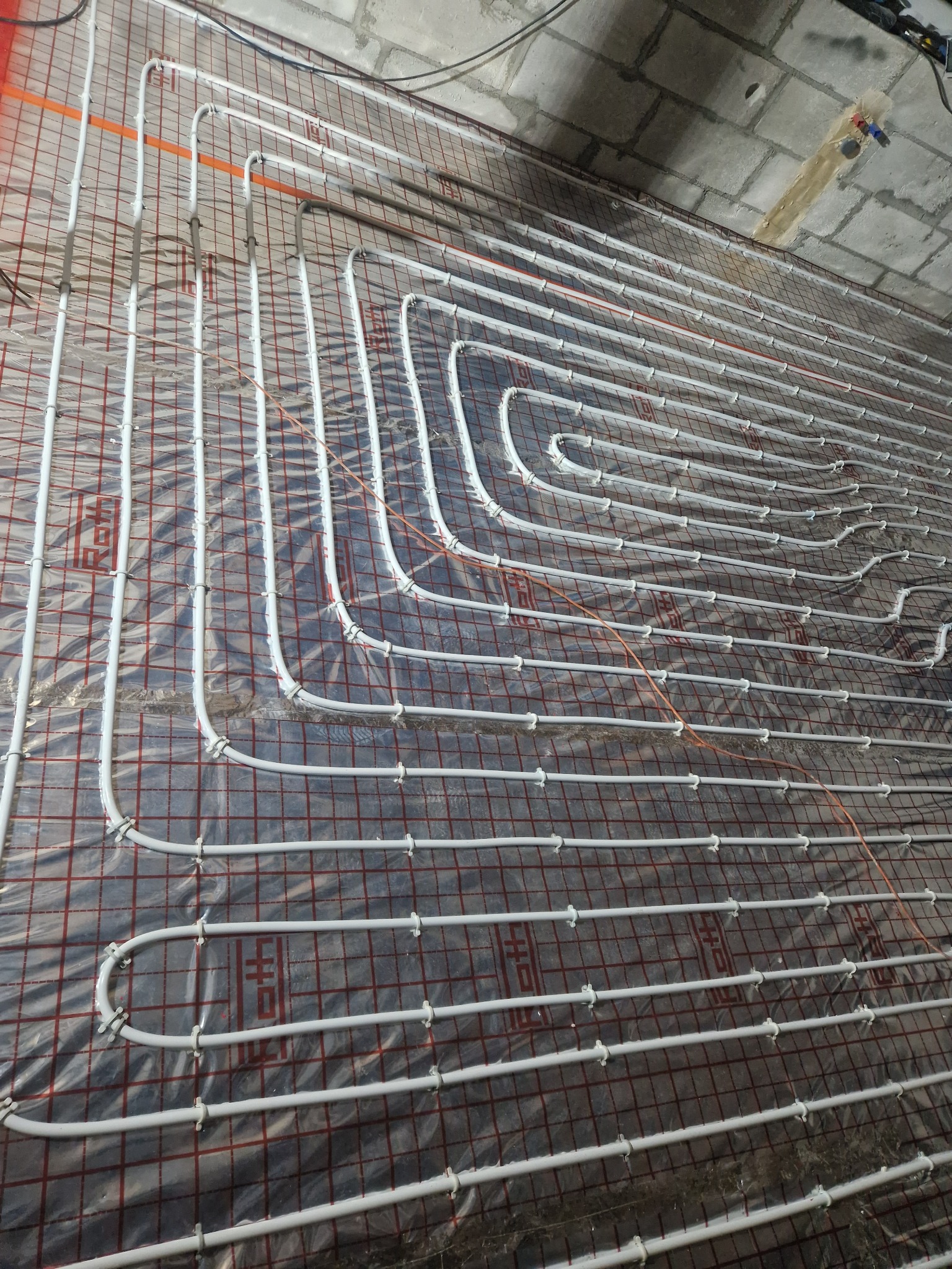 Białe rury ogrzewania podłogowego ułożone na folii z czerwoną siatką, mocowane plastikowymi klipsami do siatki, surowy betonowy sufit w tle.