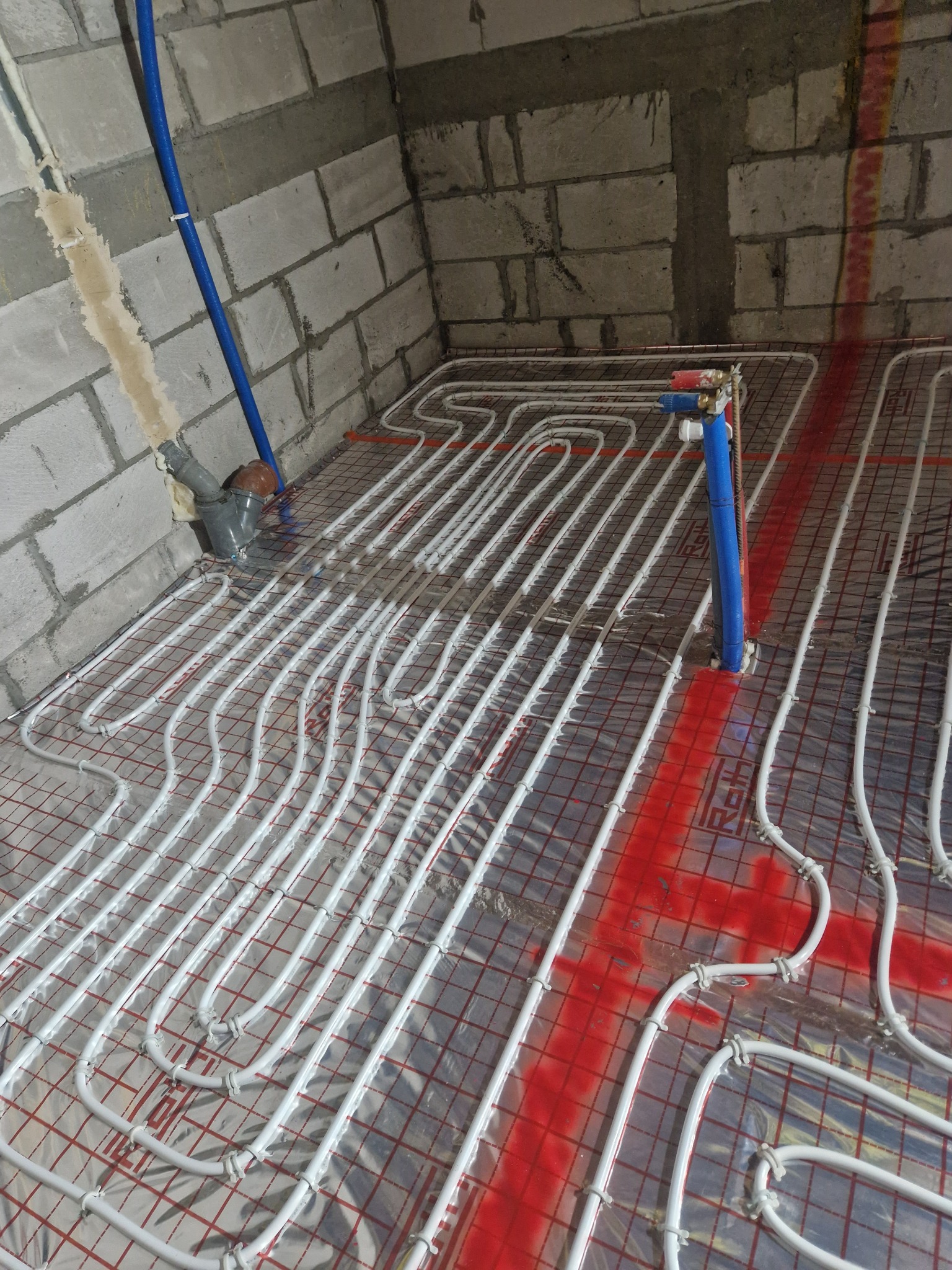 Białe rurki ogrzewania podłogowego ułożone na czerwonej siatce w surowym pomieszczeniu z betonowymi ścianami, widoczne niebieskie rury doprowadzające wodę.