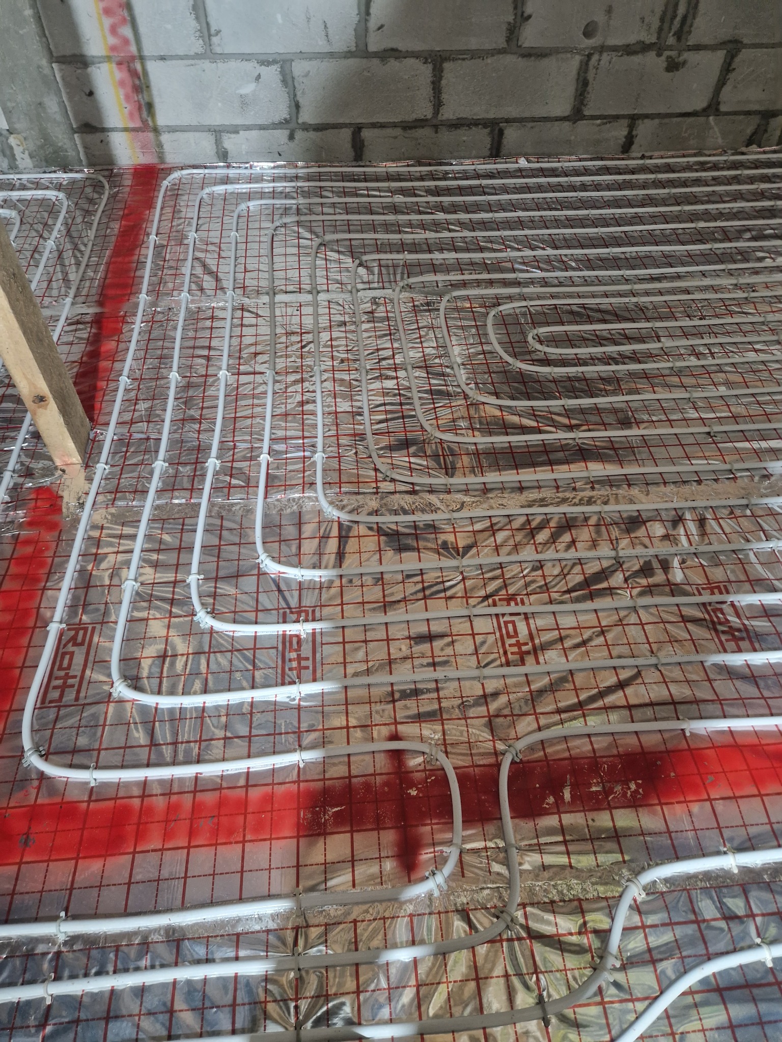 Białe rury ogrzewania podłogowego ułożone na folii izolacyjnej z czerwoną siatką, widoczne surowe ściany z betonu komórkowego i fragment drewnianej belki.
