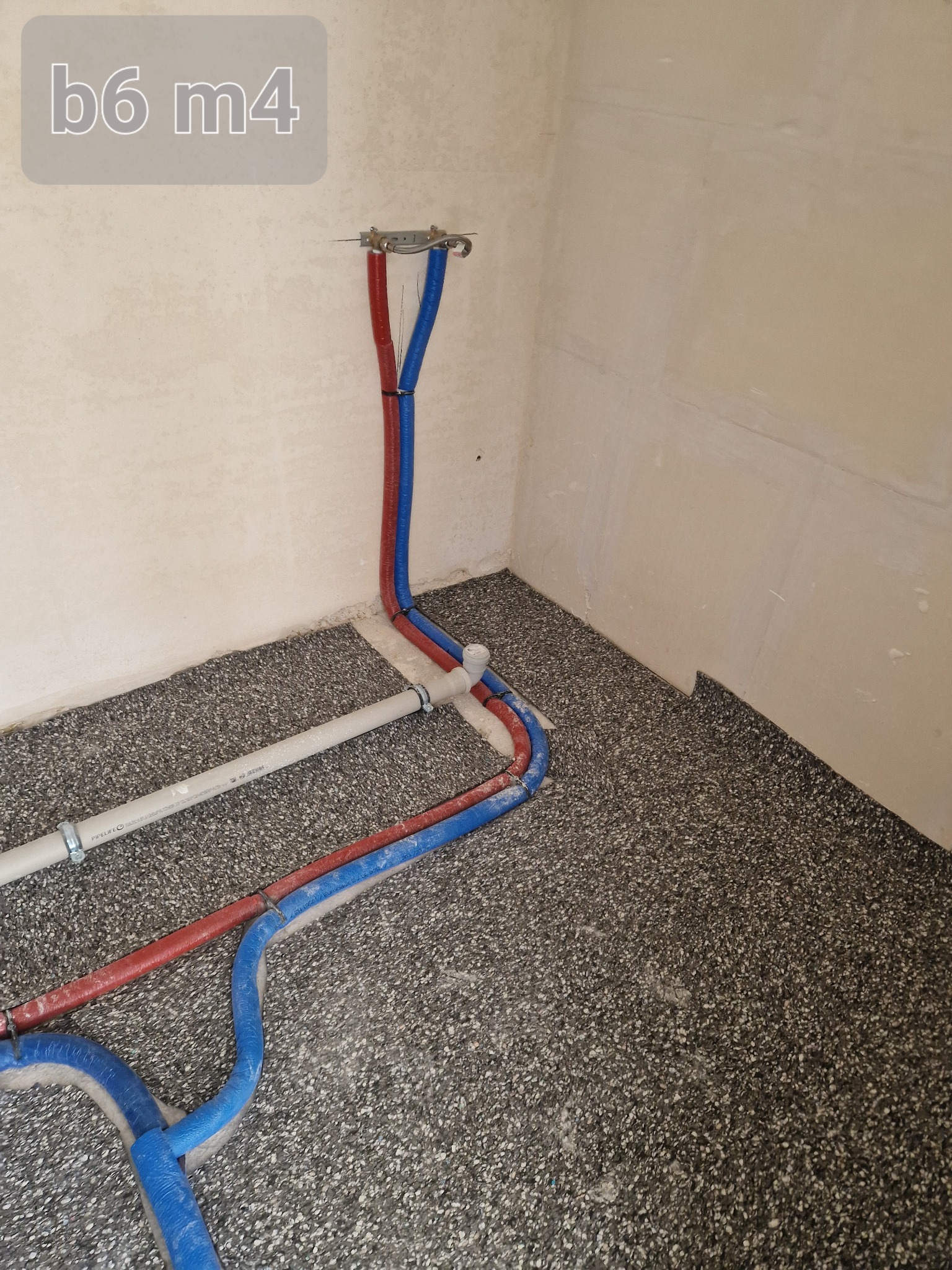 Nowe rury hydrauliczne w kolorze niebieskim i czerwonym prowadzone wzdłuż podłogi z szorstką powierzchnią, z widocznym fragmentem białej rury PCV, ściany w stanie surowym.