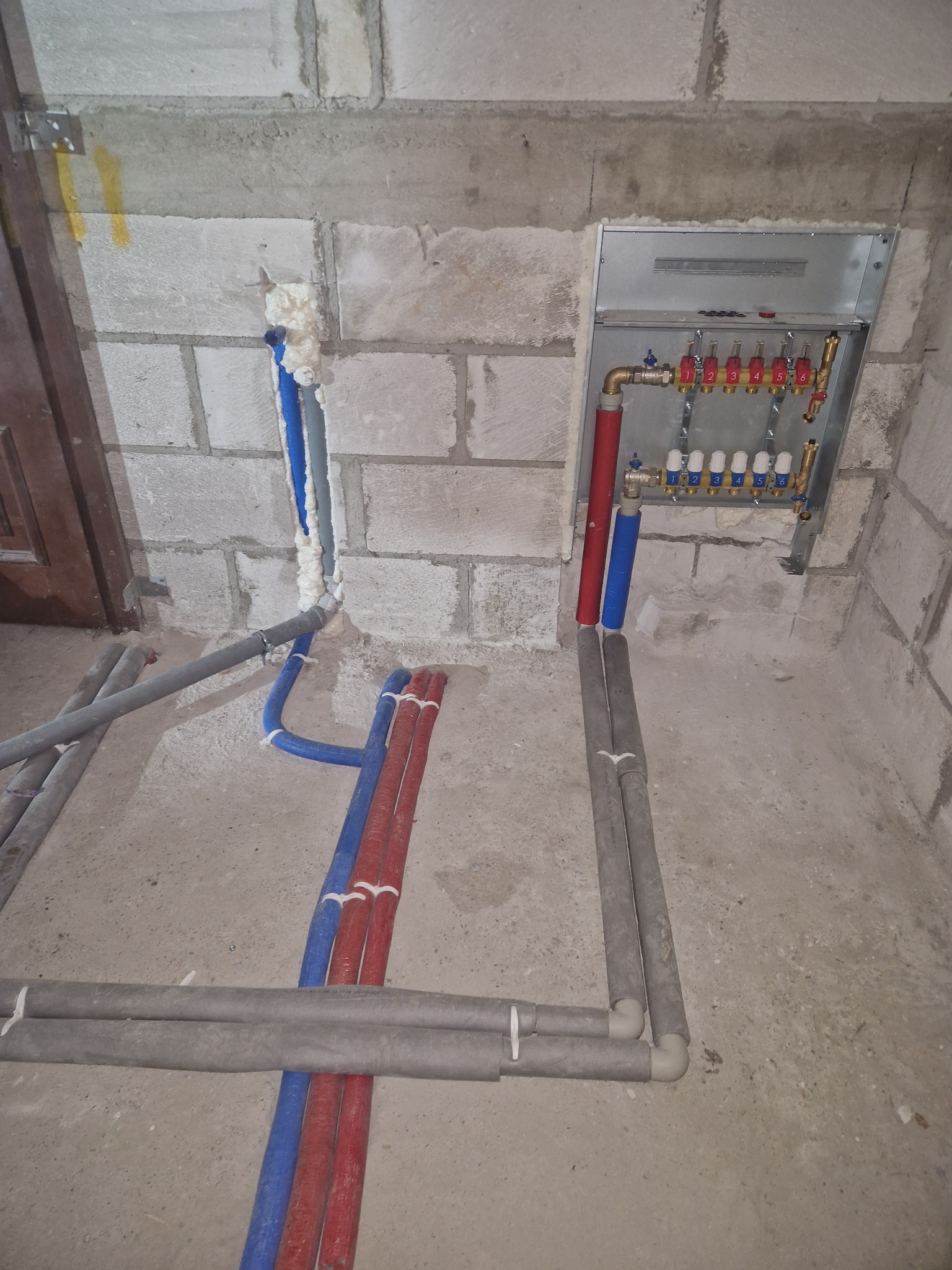 Rozdzielacz ogrzewania podłogowego z rurami w izolacji, zamontowany w surowym pomieszczeniu z betonowymi ścianami i podłogą. Widoczne rury doprowadzające i odprowadzające wodę, zabezpieczone pianką...