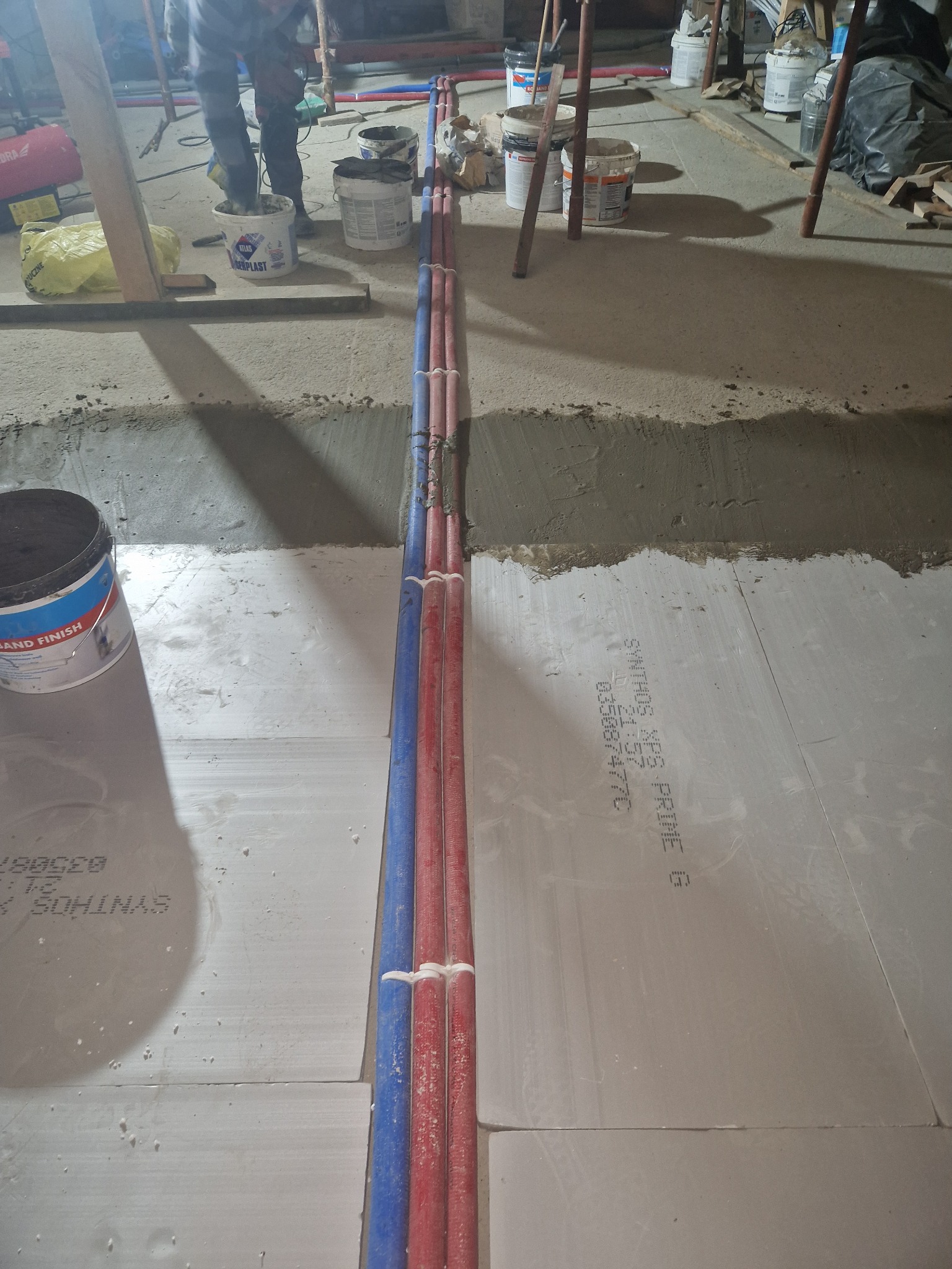 Instalacja podłogowa: rury grzewcze, niebieskie i czerwone, ułożone w wykopie na izolacji podłogowej, widoczne narzędzia i wiaderka z zaprawą w tle.