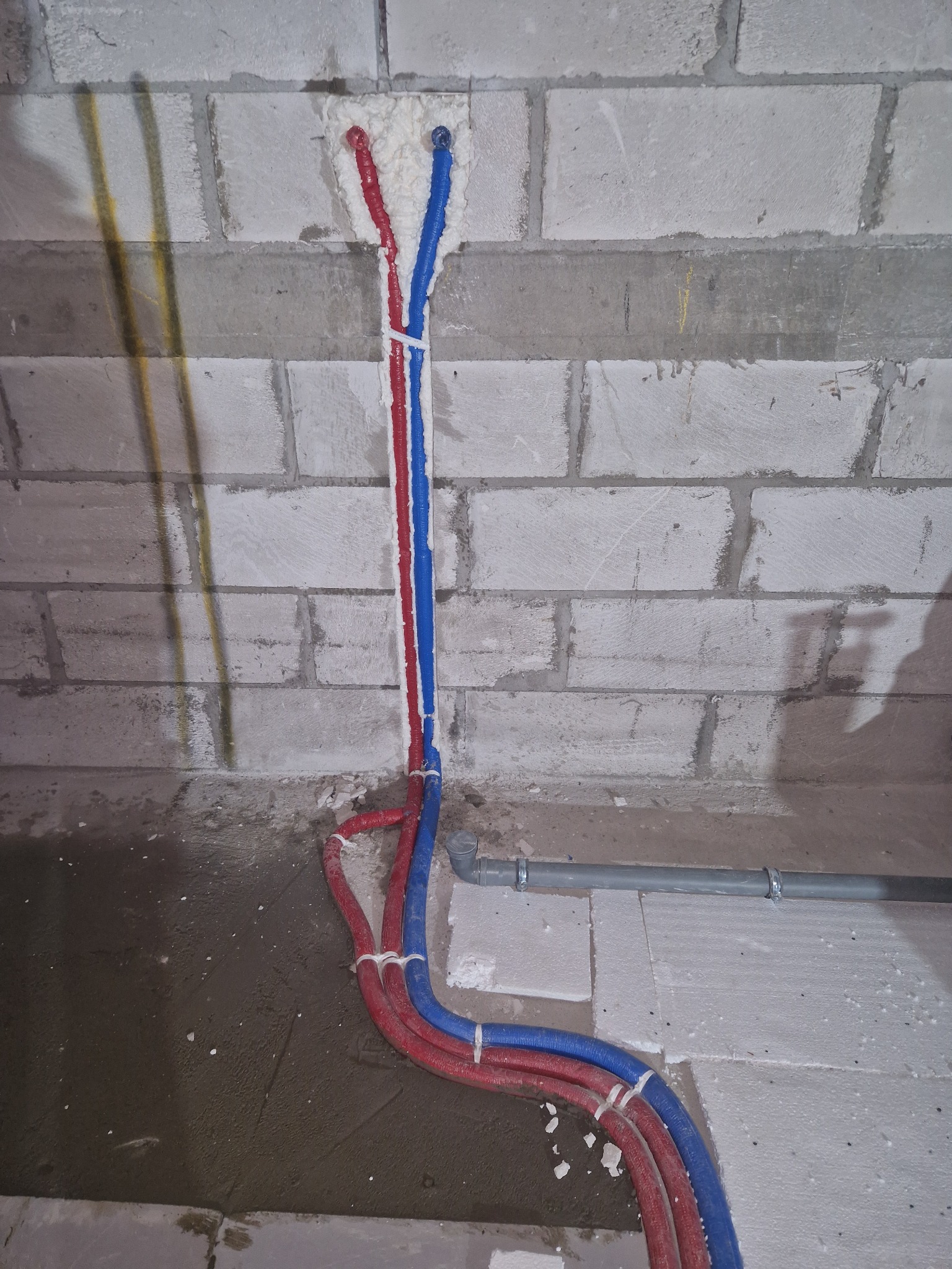 Nowe czerwono-niebieskie rury hydrauliczne prowadzone w ścianie z betonu komórkowego, zabezpieczone pianką montażową i uchwytami, widok z dołu na surowym betonie.