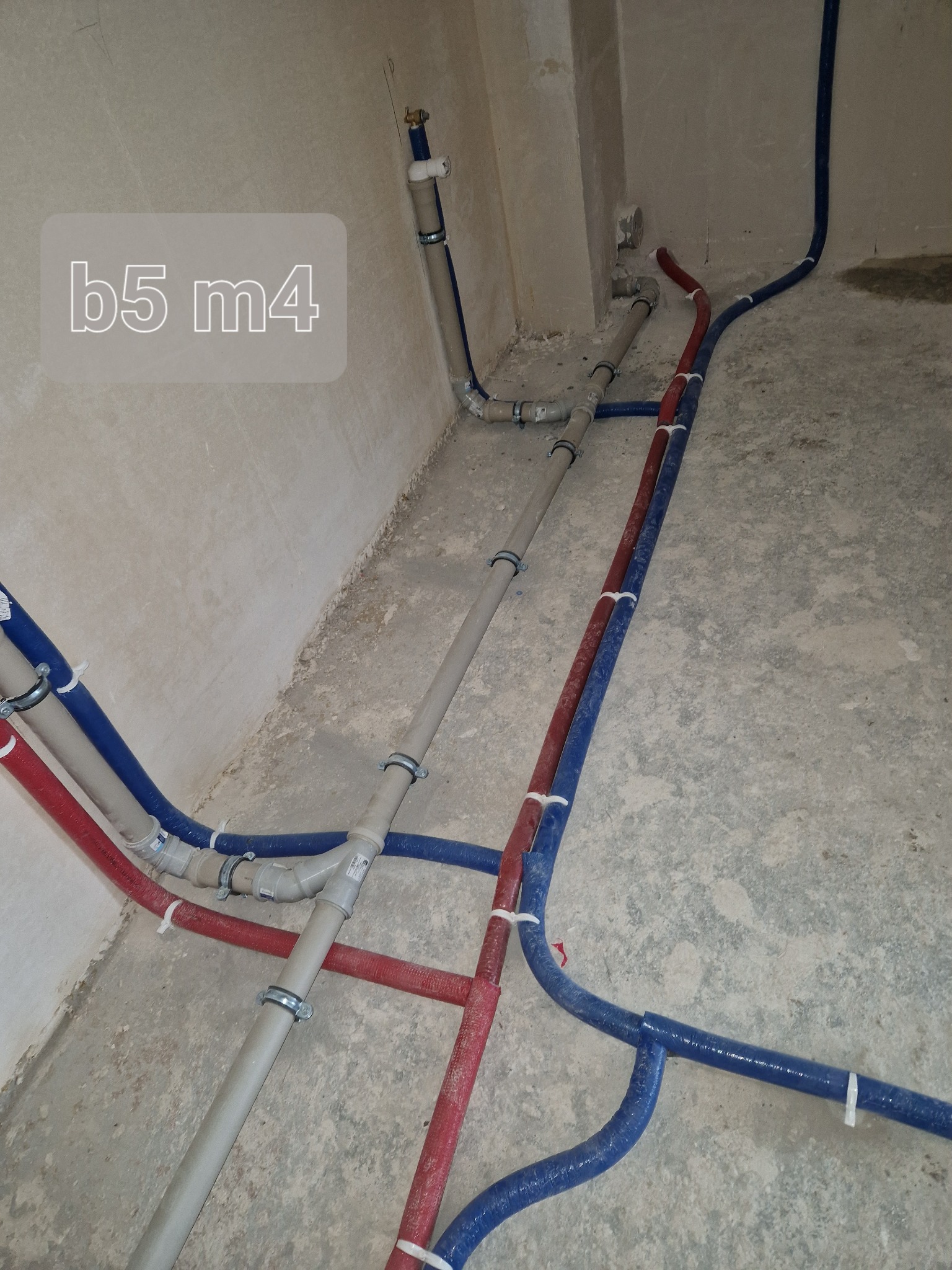 Nowe rury wodne i grzewcze, czerwone i niebieskie, prowadzone wzdłuż ściany i podłogi, zabezpieczone białymi opaskami zaciskowymi, w surowym wnętrzu.