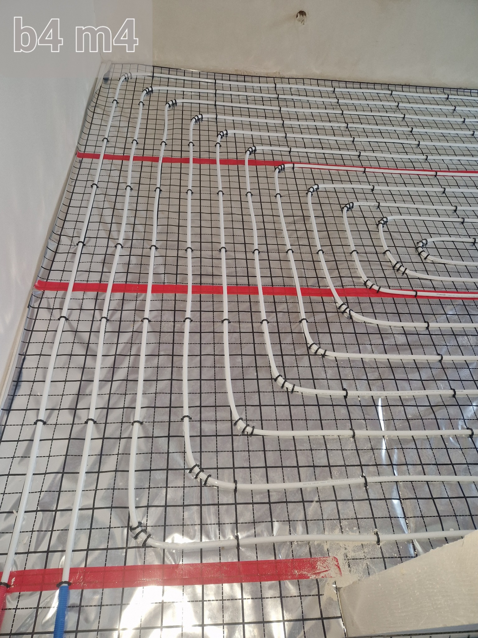 Białe rurki ogrzewania podłogowego, mocowane czarnymi klipsami do folii z nadrukowaną siatką, ułożone na izolacji ze srebrną folią aluminiową, z widocznymi czerwonymi pasami taśmy mocującej, przed...