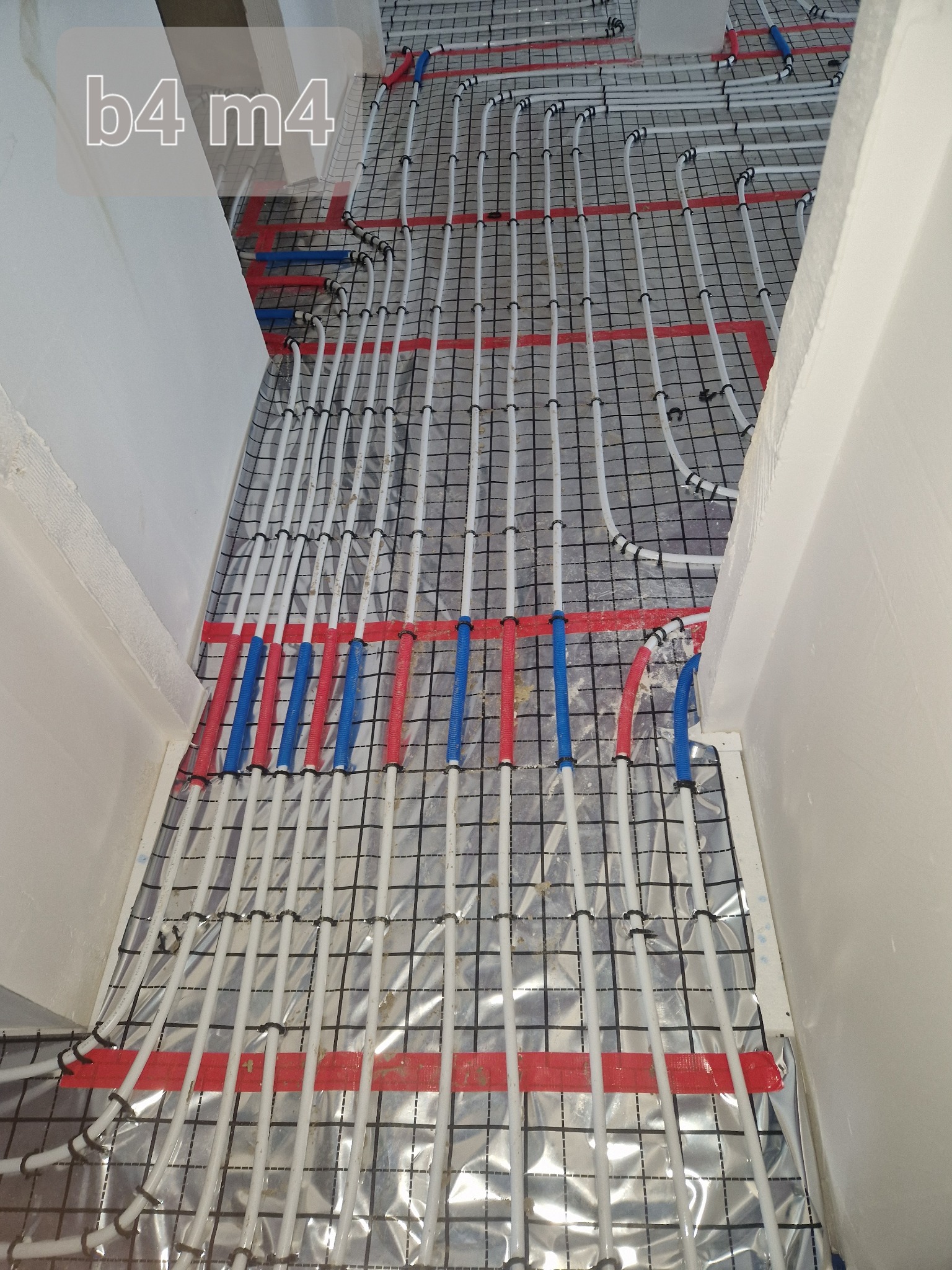 Rozłożone białe rury ogrzewania podłogowego z czerwonymi i niebieskimi oznaczeniami na czarnej siatce montażowej, widok z góry w surowym stanie.