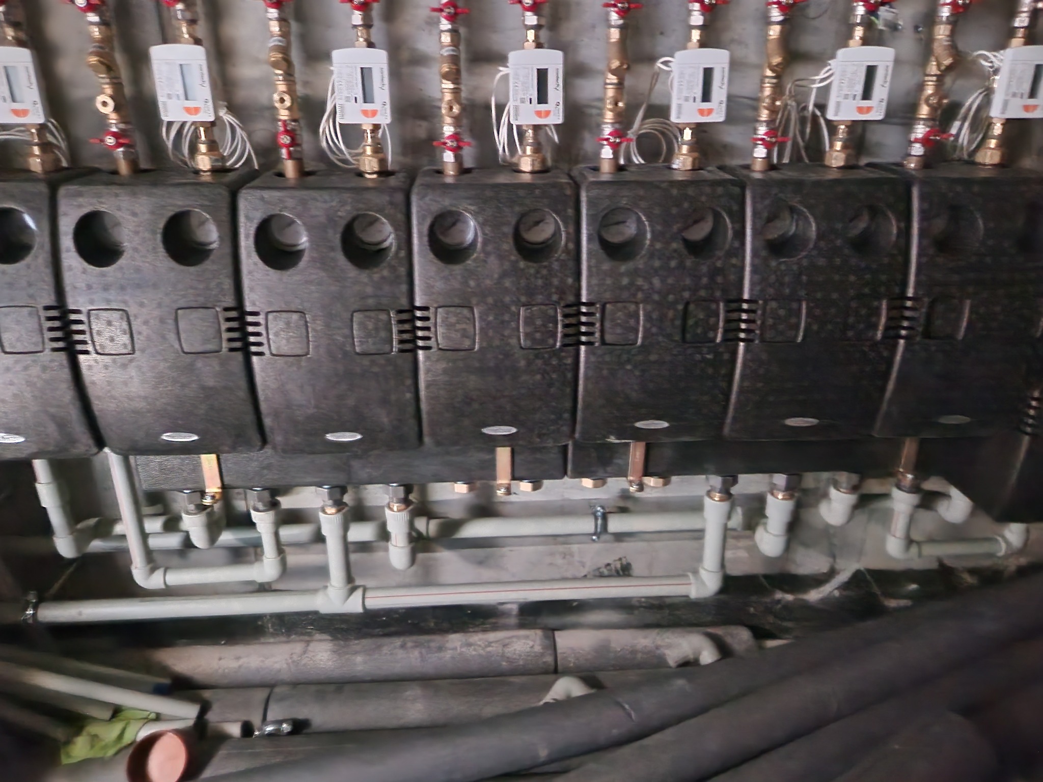 Rozdzielacz centralnego ogrzewania z licznikami ciepła, widoczne rury i zawory, szare tło ściany.