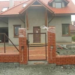 Zyg-Mal usługi remontowo budowlane - Surowy stan elewacji domu jednorodzinnego z widoczną konstrukcją tarasu, murowanymi słupkami ogrodzenia z cegły klinkierowej i metalową furtką w trakcie montażu, na pierwszym planie rozsypany piasek.