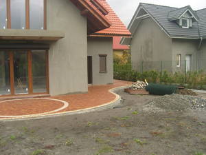 Kaskadowy chodnik z czerwonej kostki brukowej, obrzeża z jasnej kostki, świeżo ułożony na tle surowego ogrodu i dwóch domów w szarym tynku.