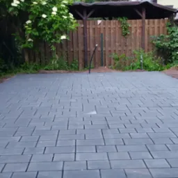 Szara kostka brukowa ułożona na posesji, widoczna zieleń i drewniana altana w tle. Regularny wzór kostki, świeżo zakończone prace.